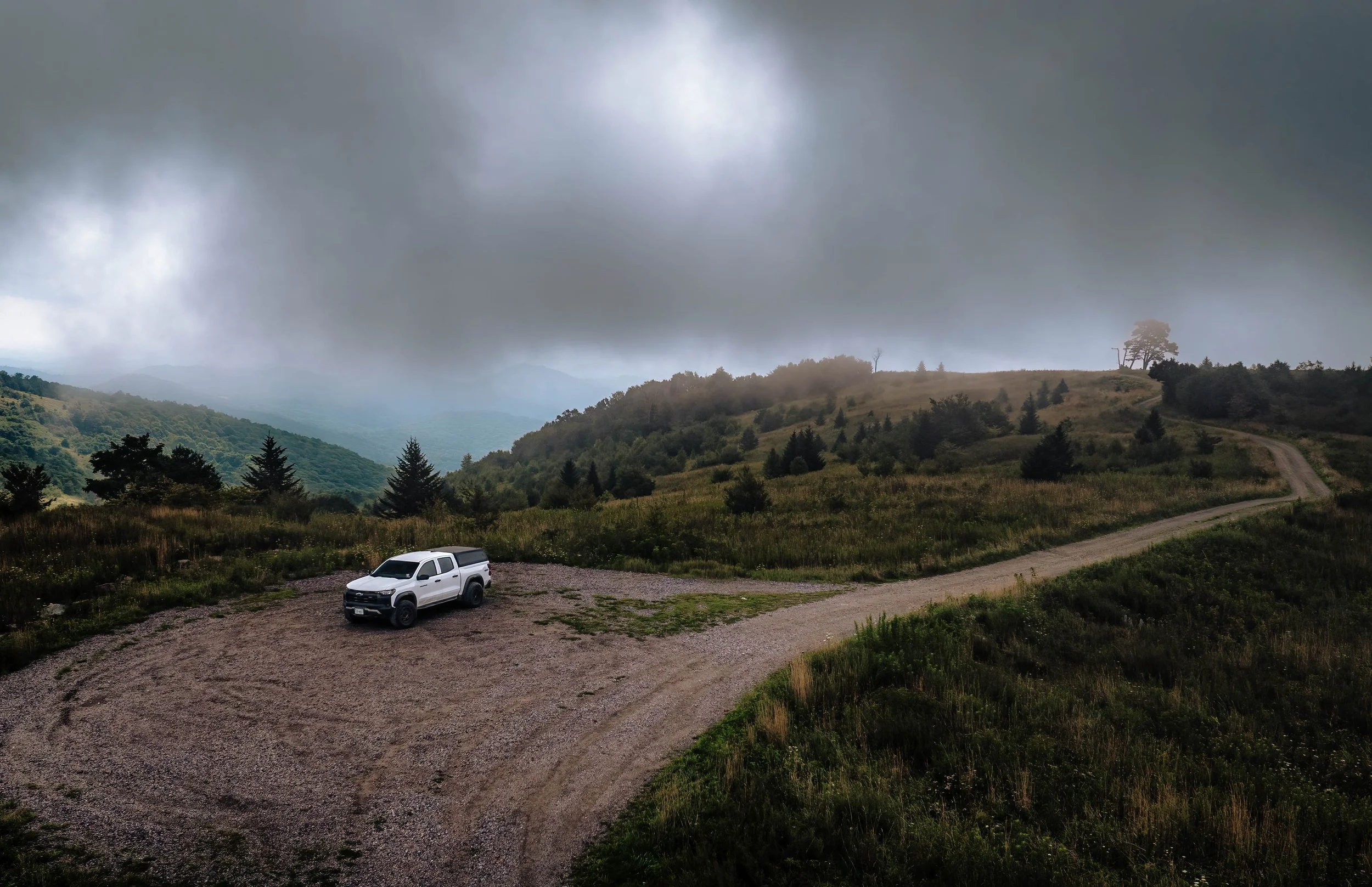 A white pickup truck parked on a dirt road in a hilly, rural area with lush green trees and grass. Foreboding dark, cloudy sky overhead.