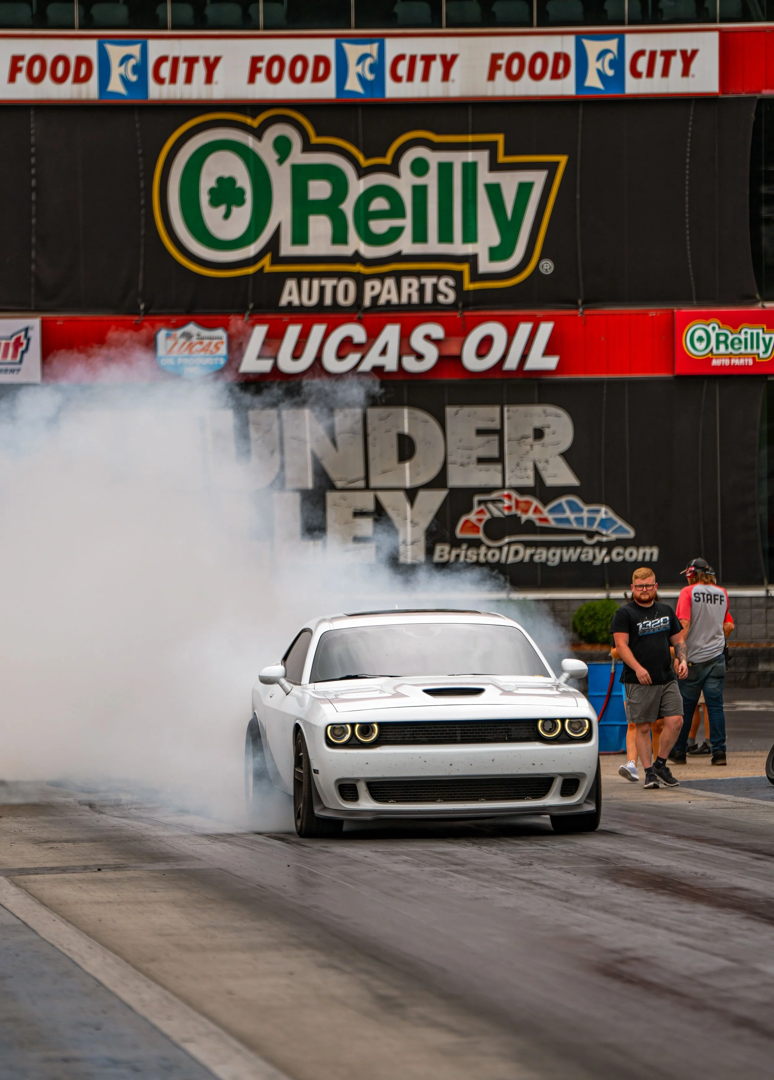 A white muscle car performing a burnout at Bristol Dragway, with tire smoke billowing behind it, during a racing event. Background features banners for O'Reilly Auto Parts, Lucas Oil, and Under Ty.