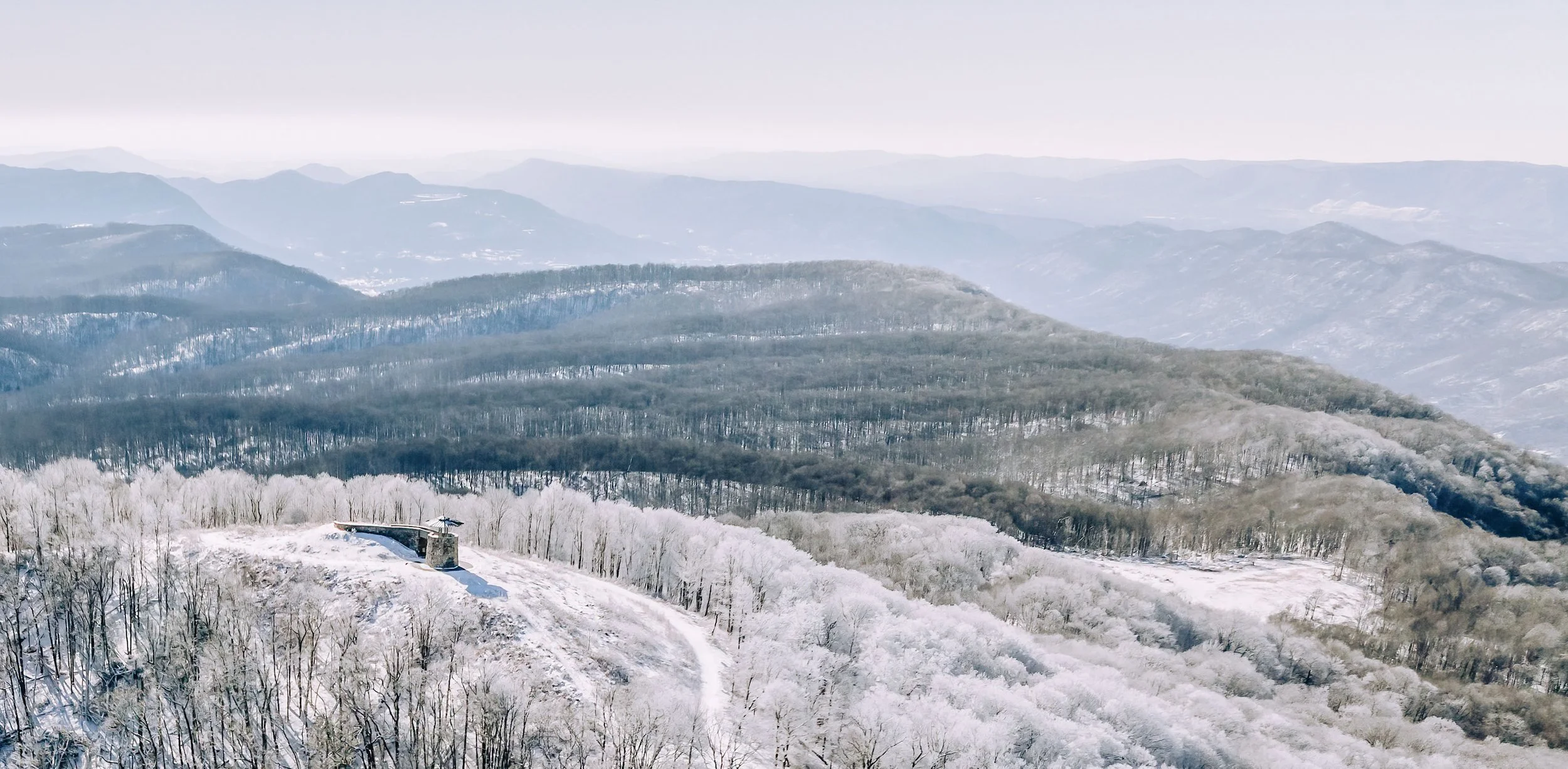 A snowy mountain landscape with layers of forested hills receding into the distance and a small building or observation station on a snowy hill in the foreground.