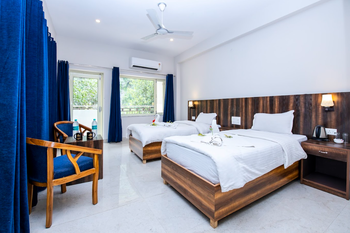Hotel room with two single beds, white linens, wooden headboards, blue curtains, a small dining table with two bottled waters, and a window with a view of green trees.