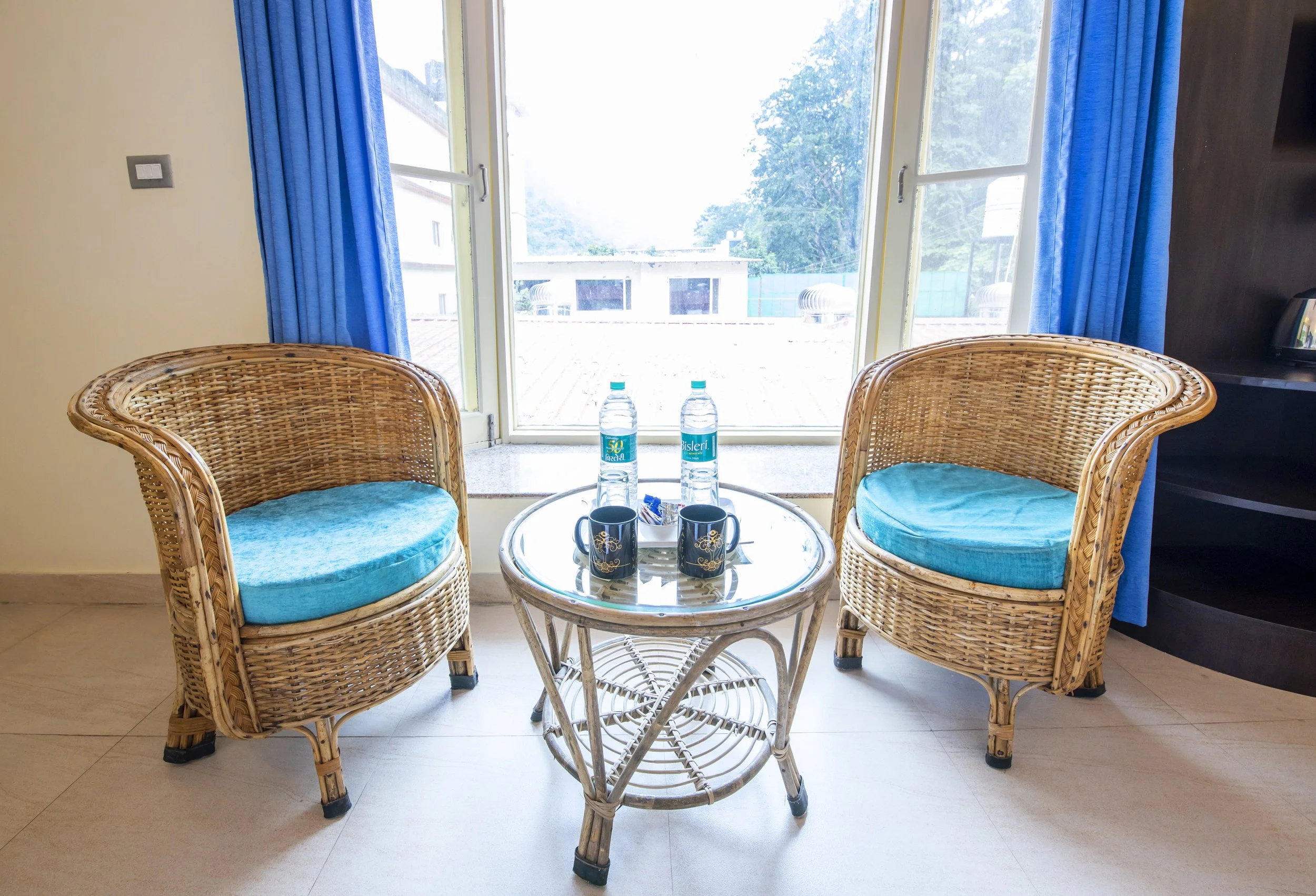 Two wicker chairs with blue cushions facing a glass-top table with two bottles of water, two black coffee mugs, and a small tray, in front of a large window with blue curtains.