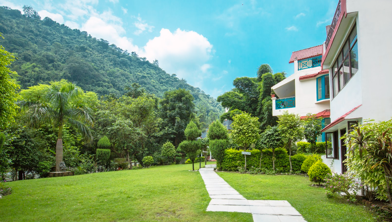 A lush green garden with neatly trimmed bushes, trees, and a paved walkway leading towards mountains in the background. A white building with blue accents and red roofs is on the right side of the image. The sky is blue with some clouds.