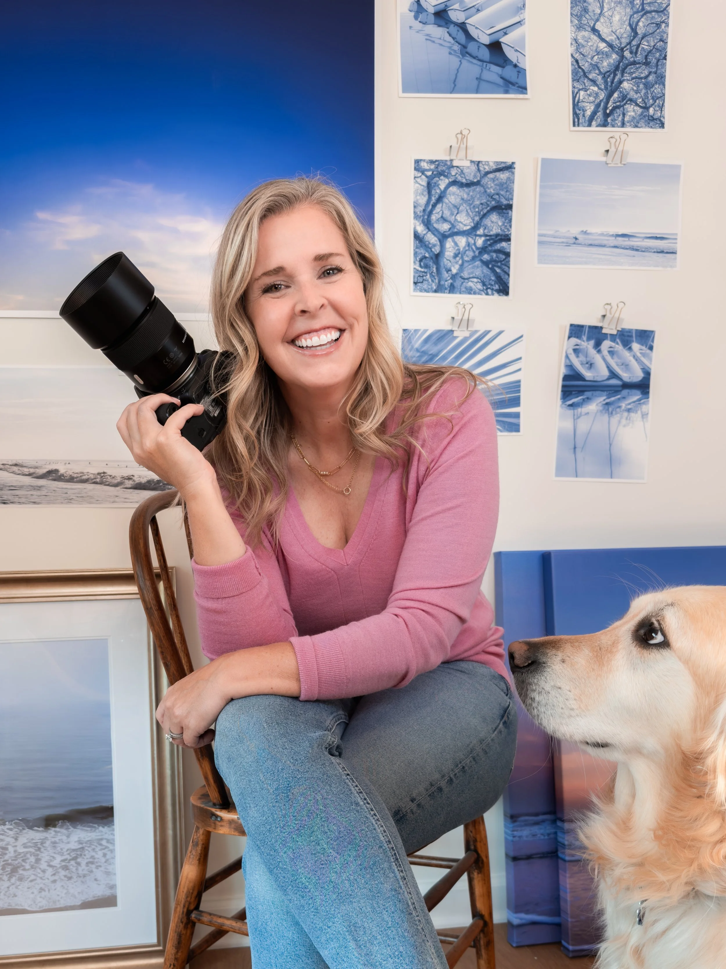 A woman with blonde hair smiling and holding a camera while sitting on a wooden chair, with a golden retriever looking at her, in front of a wall decorated with blue-themed artwork and photographs.