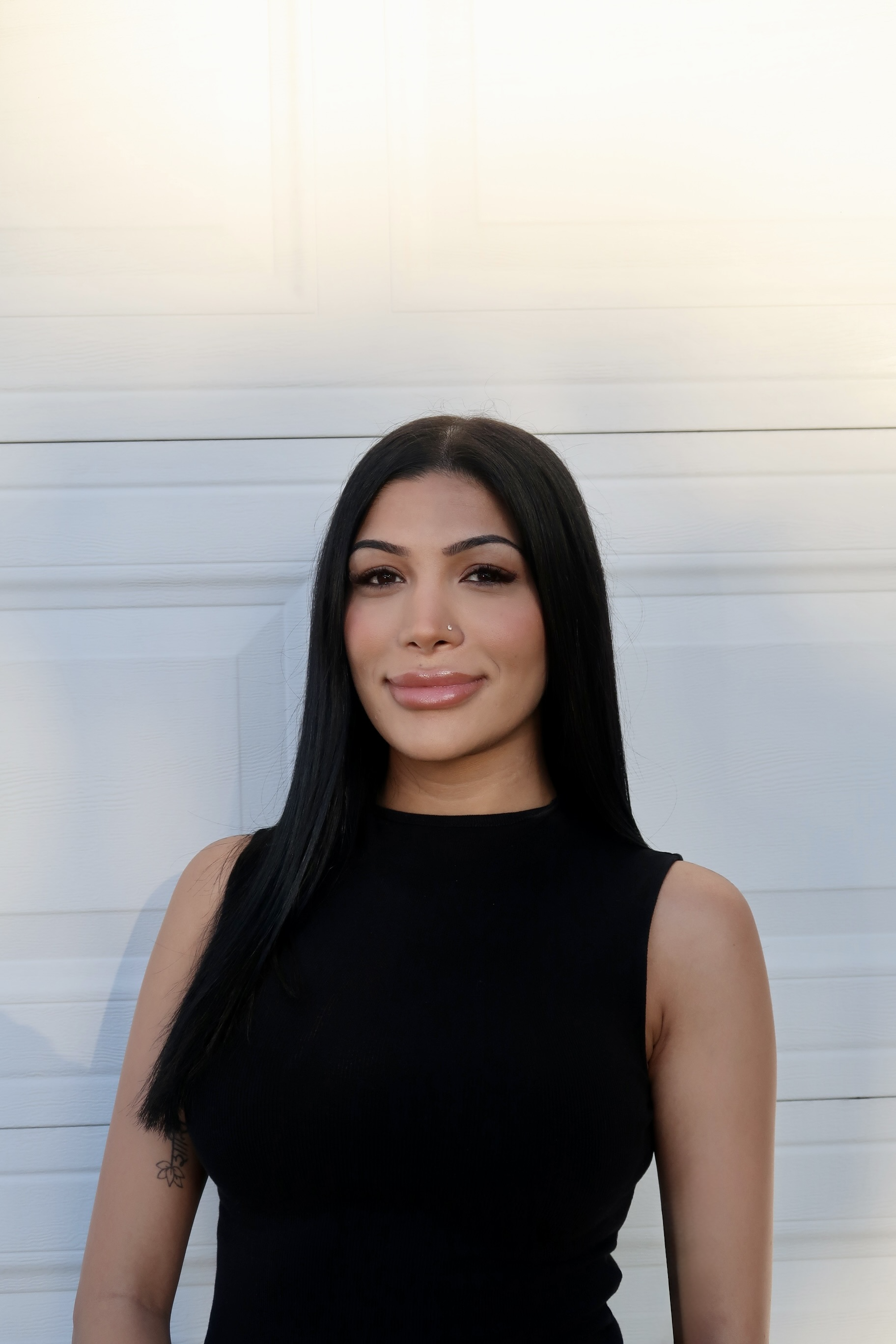 A young woman with long black hair, wearing a sleeveless black top, smiling softly, standing outdoors in front of a white garage door.