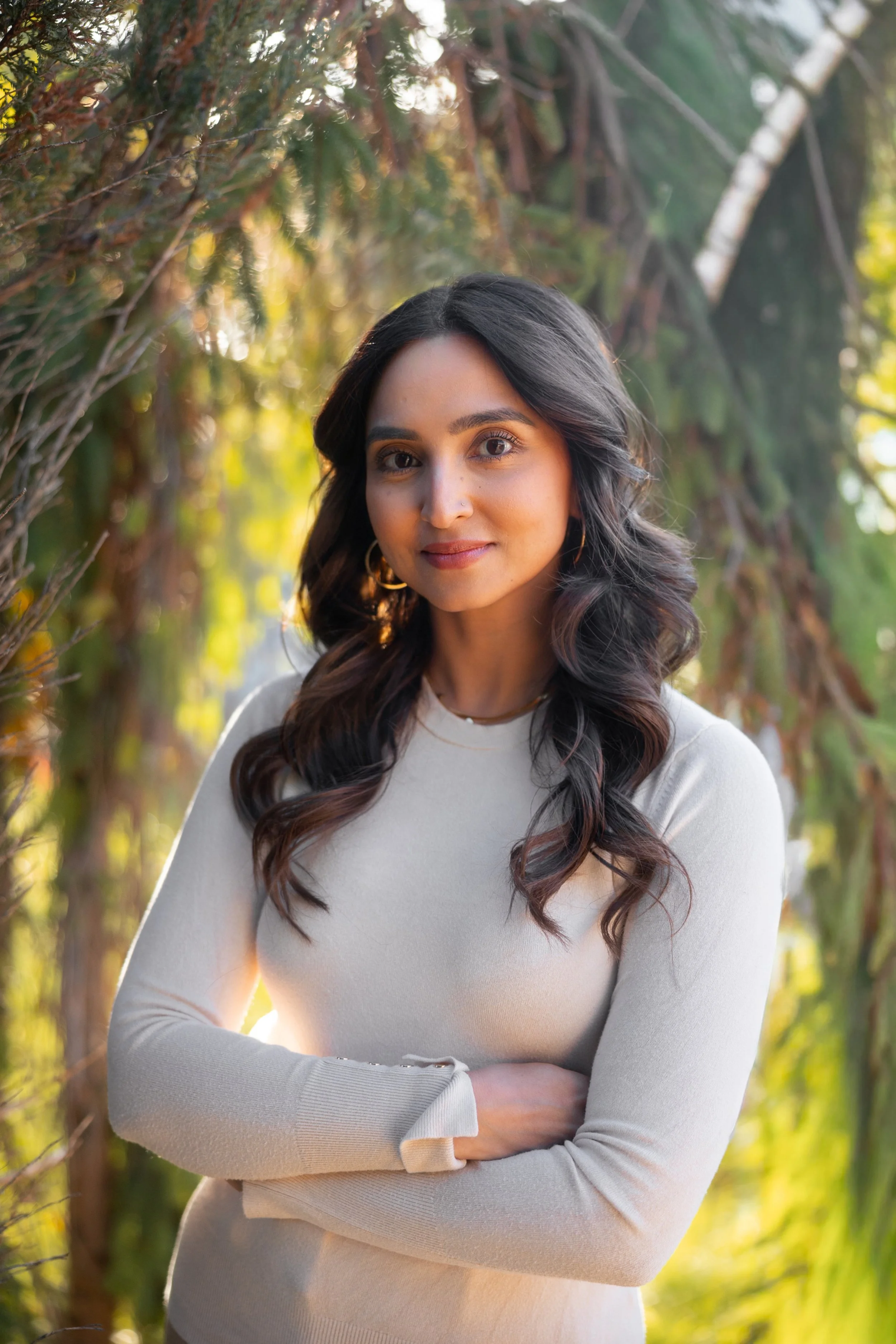A woman with long, dark, wavy hair wearing a light-colored sweater, standing outdoors with her arms crossed and a gentle smile, surrounded by green and yellow foliage.
