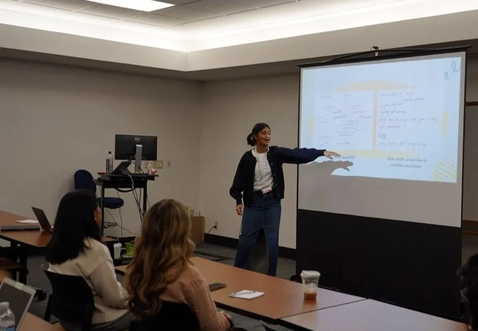 A woman is giving a presentation to a group of three women in a conference room. She is pointing at a projection screen displaying handwritten notes.