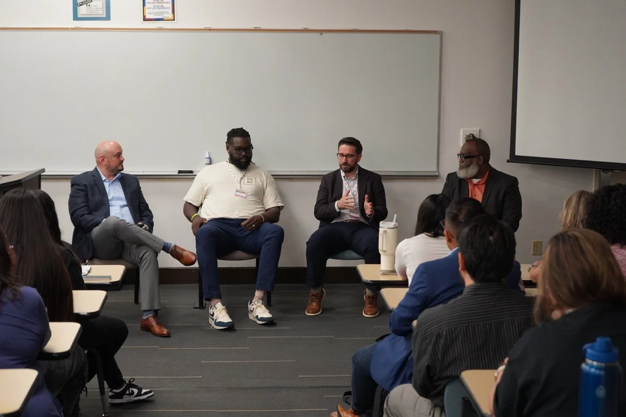 A panel discussion in a classroom with four men seated at the front, speaking to an audience of students. The classroom has a whiteboard, a projector screen, and pictures on the wall.