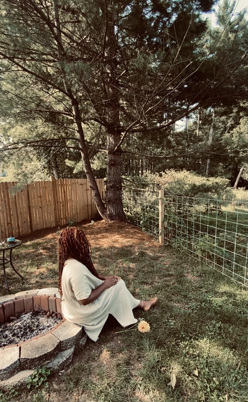 Woman in contemplation with long dreadlocks sitting on the edge of a fire pit in a backyard, facing a large tree with a fence and a wire fence in the background, during daytime. Self-portrait photographed by udee bassey.