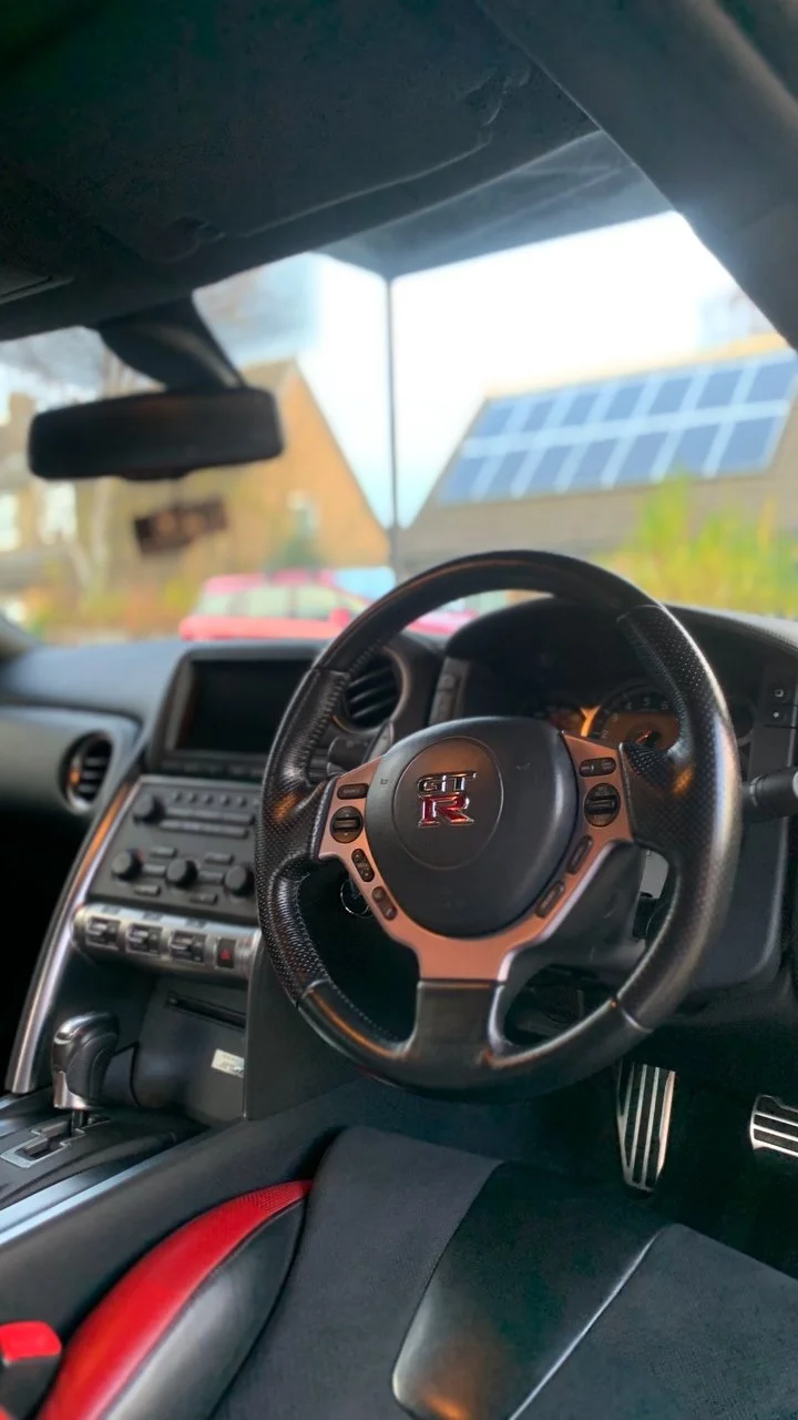 Interior of a Nissan GT-R with a steering wheel, dashboard, and gear shift. The steering wheel features the GT-R logo, and there's a view of houses with solar panels on the roofs through the windshield.