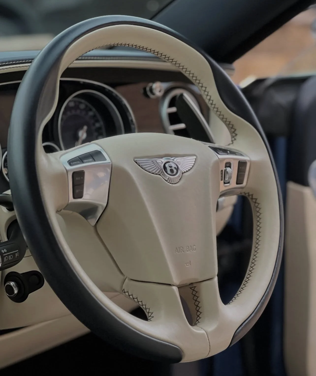 Inside view of a luxury car's steering wheel, featuring the Bentley logo, with a cream-colored leather wrap with black accent and detailed stitching.