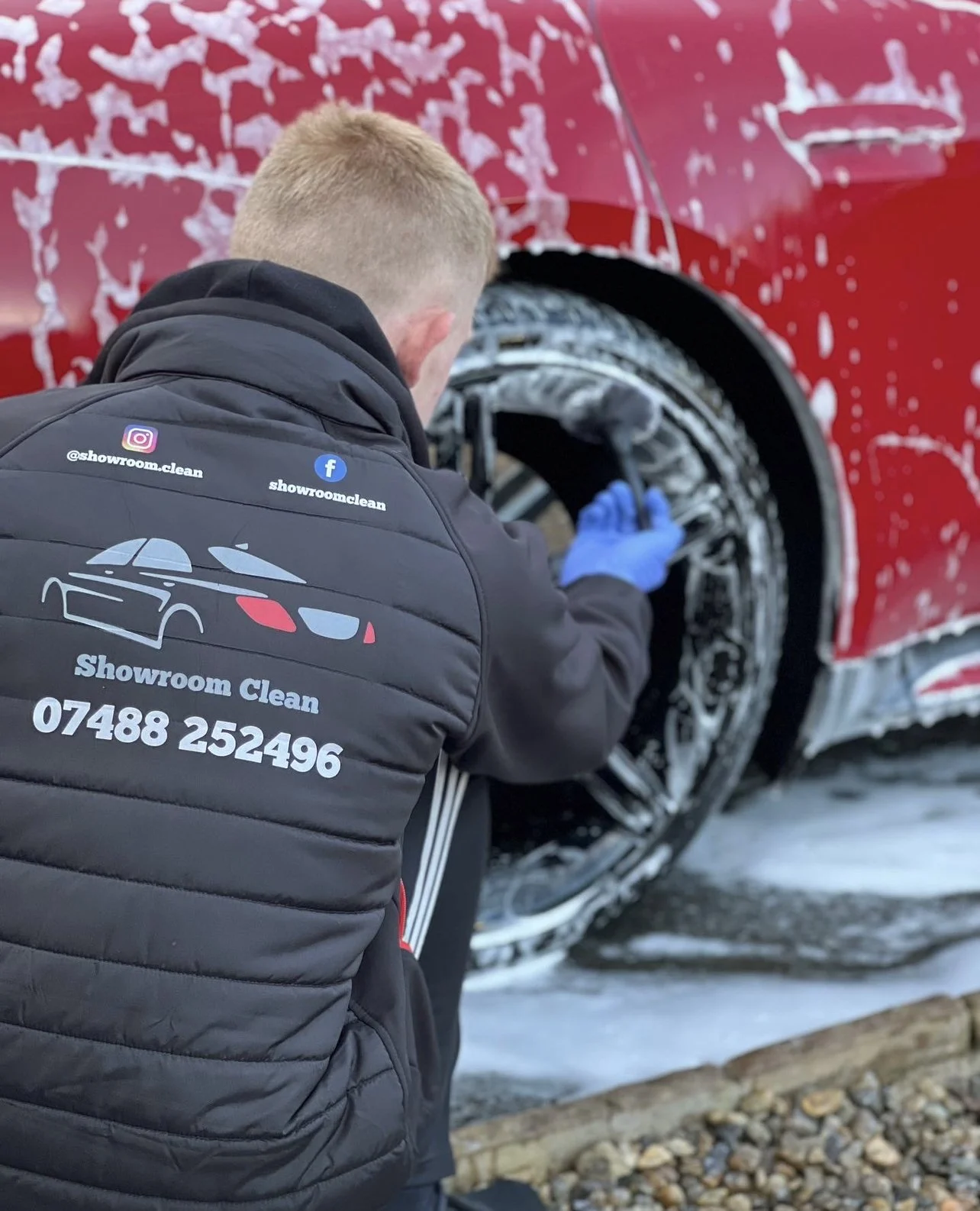A person in a Showroom Clean jacket washing a red car with soap, foam, and water on the vehicle's wheel and body.