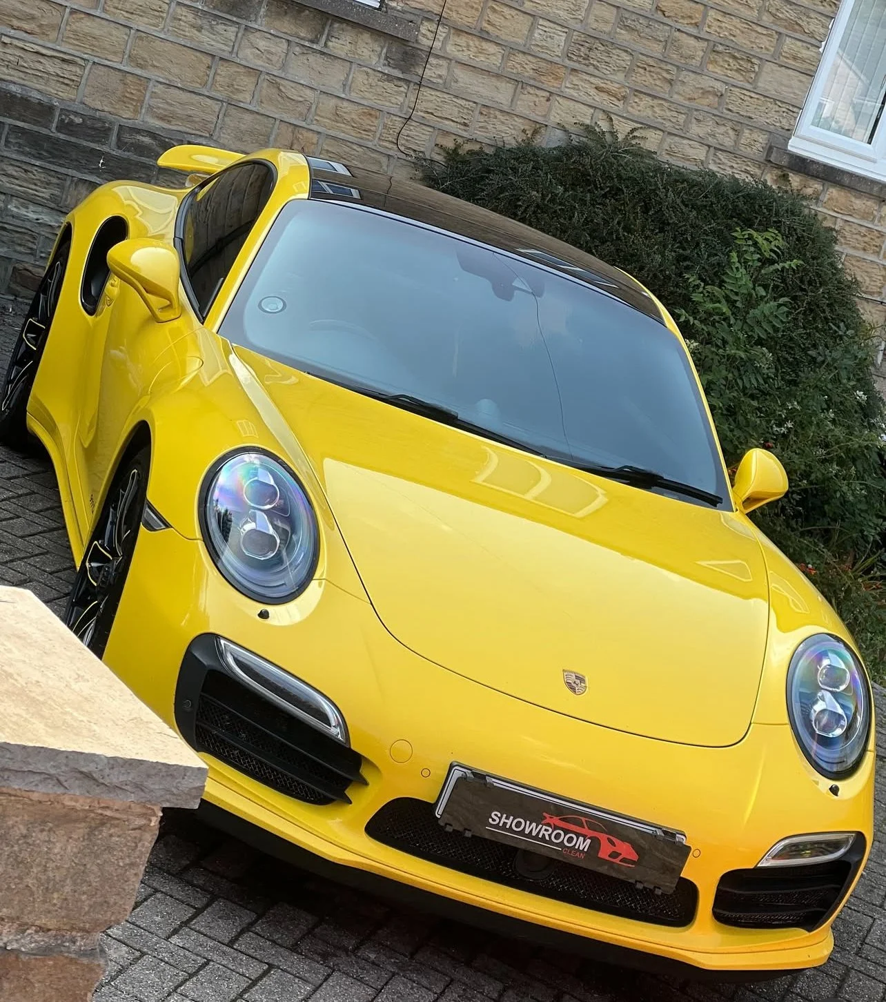 Yellow Porsche sports car parked on a driveway in front of a brick wall and green bush.
