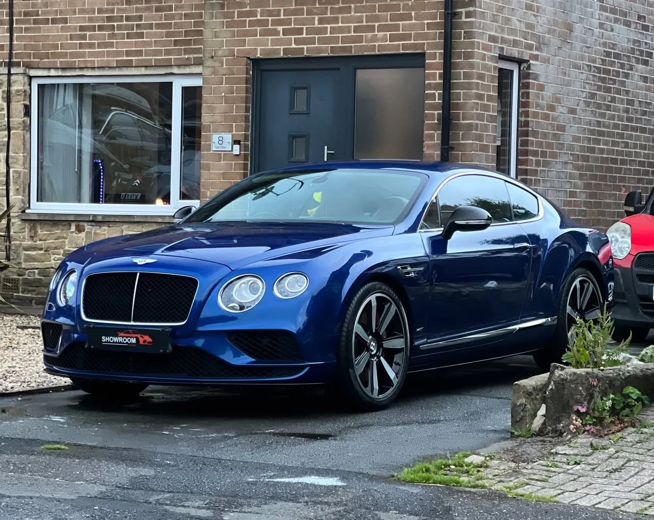 A blue luxury sports car parked on a driveway in front of a brick house with black door and window.
