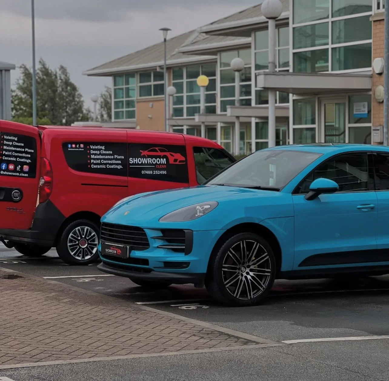 Blue car parked next to a red van in a parking lot with modern buildings in the background.