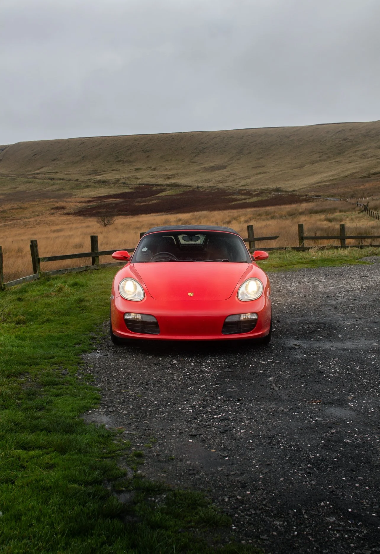 Red Porsche sports car parked on gravel driveway in a rural landscape with grassy fields, wooden fence, and rolling hills under an overcast sky.