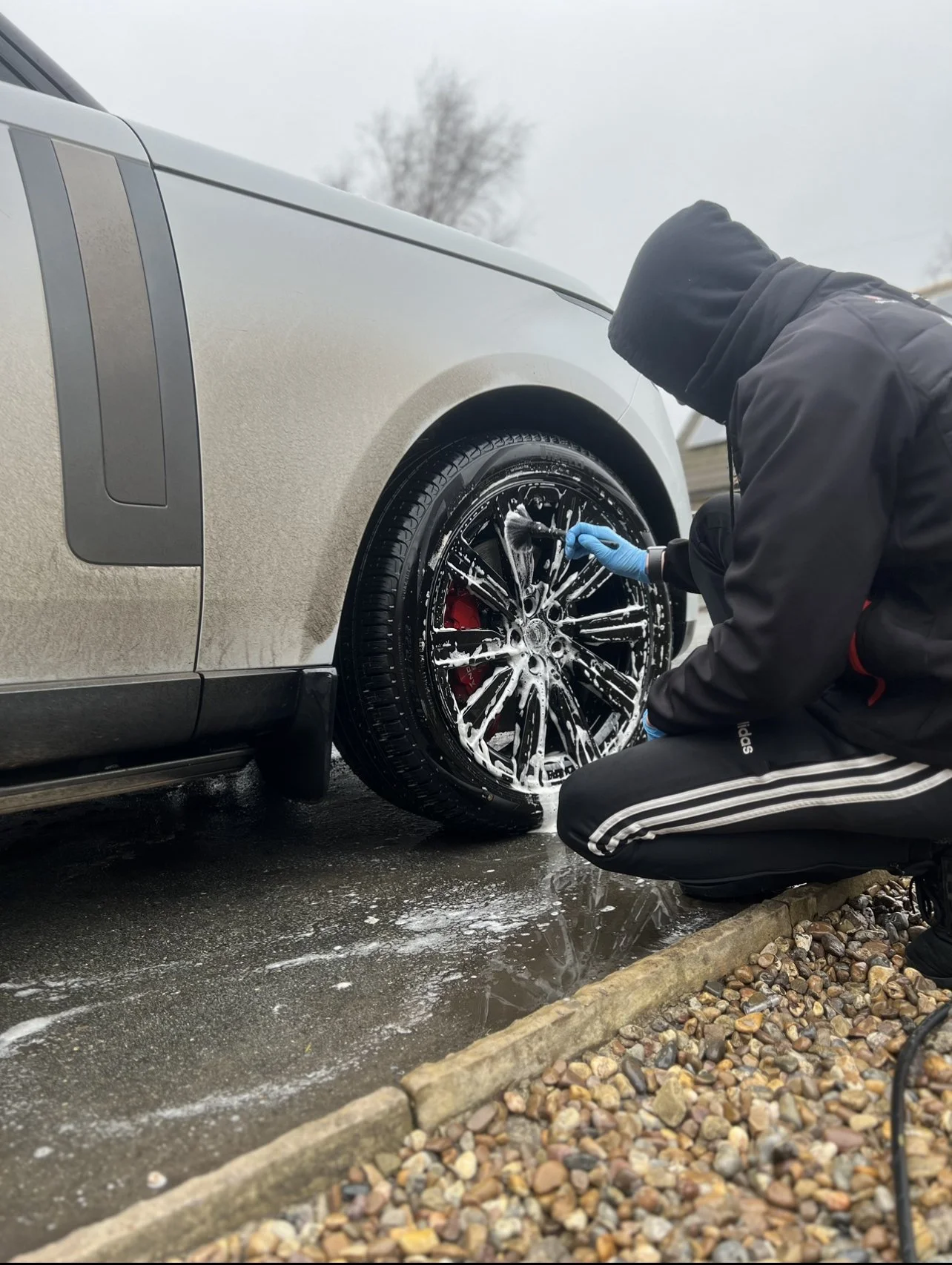 Person washing black car wheel with soap and water, wearing black hoodie, kneeling on gravel driveway.