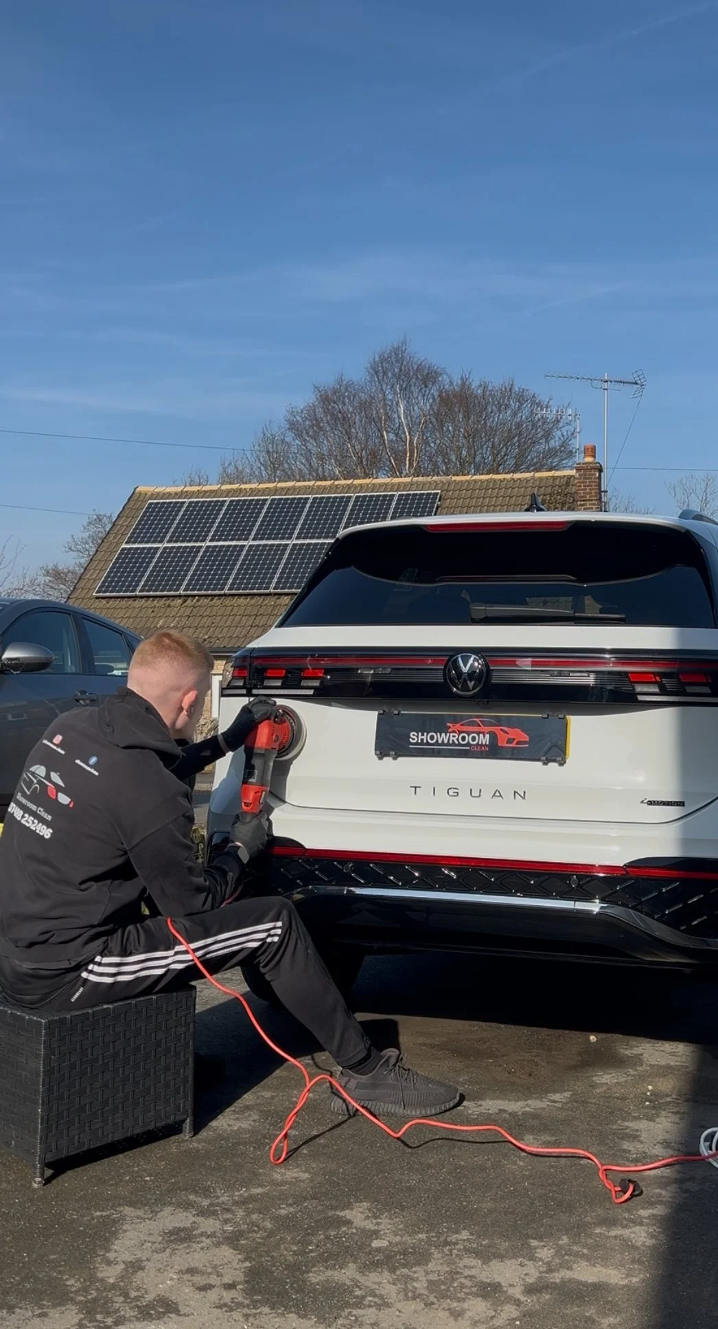 Person polishing the rear of a Volkswagen Tiguan SUV with a polishing or cleaning machine, parked outdoors on a sunny day.