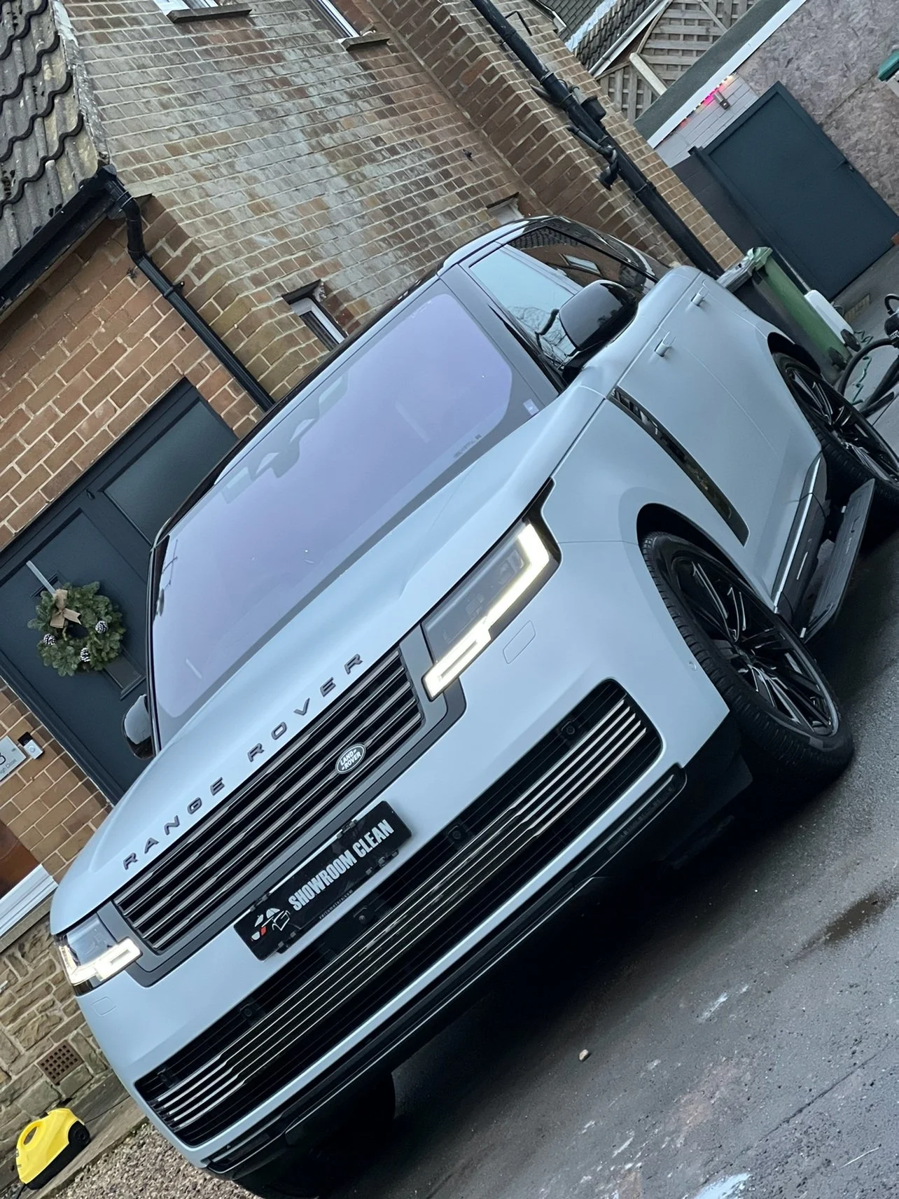 A white Range Rover parked on a narrow driveway next to a brick house, with a wreath on the front door and a yellow container on the ground nearby.