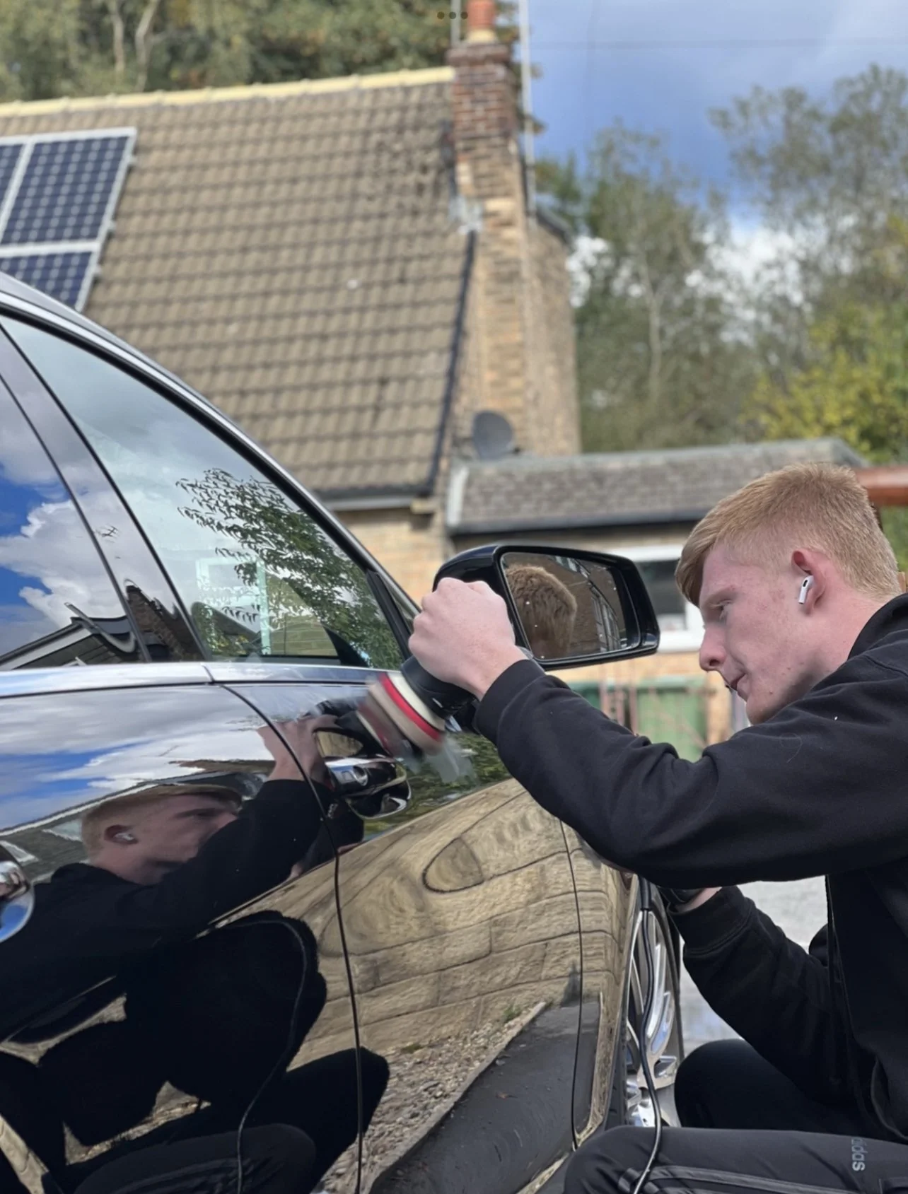 A young man with red hair and wearing wireless earbuds is polishing a black car with a battery-powered polisher, with his reflection visible on the car's shiny surface, in a residential driveway.