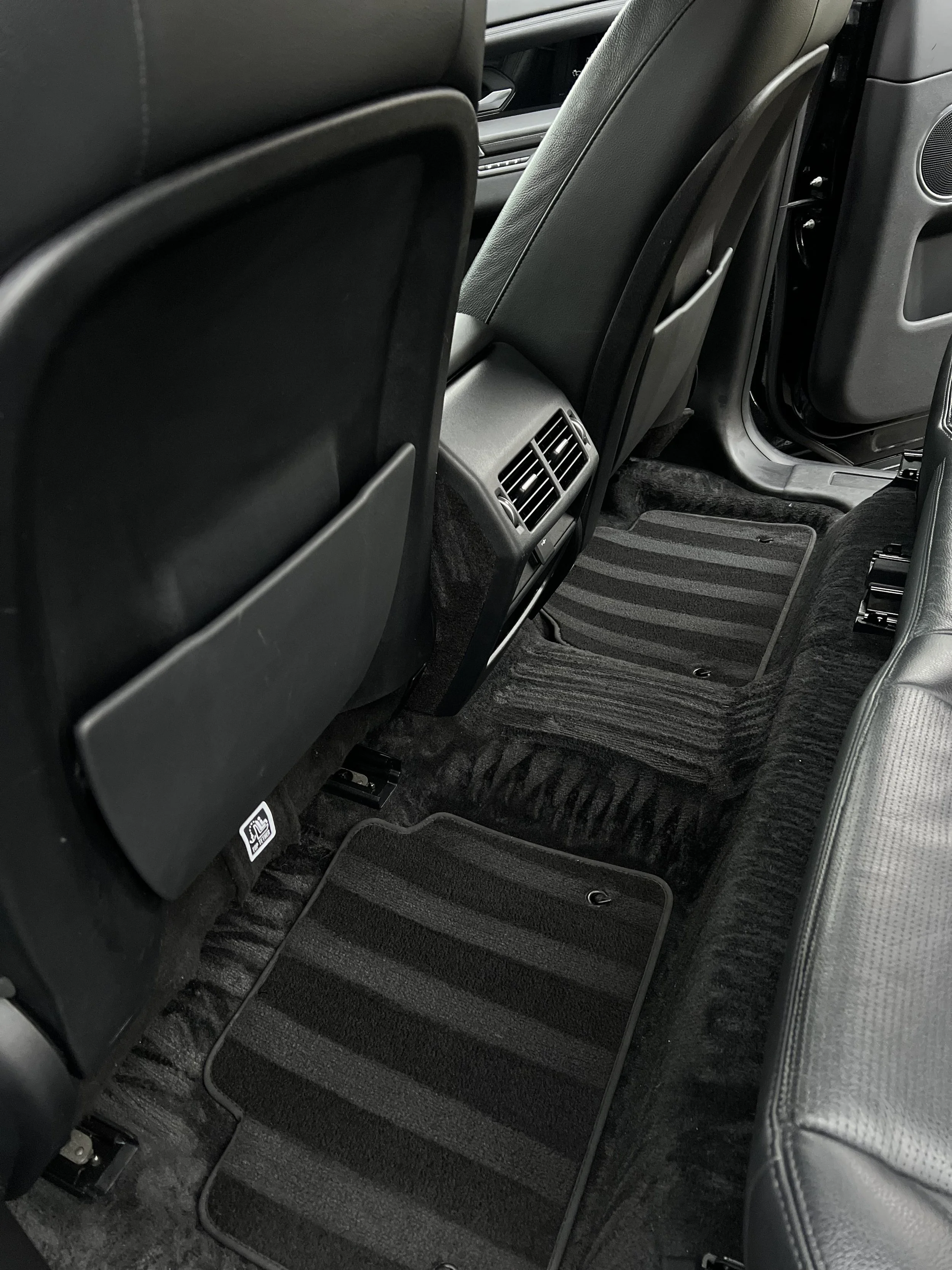 Interior of a car's back seat area showing black seats, floor mats, and rear air vents.