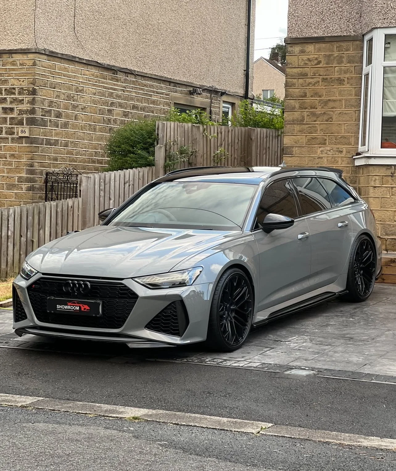 Gray Audi sedan parked on a driveway in front of a wooden fence and brick house.