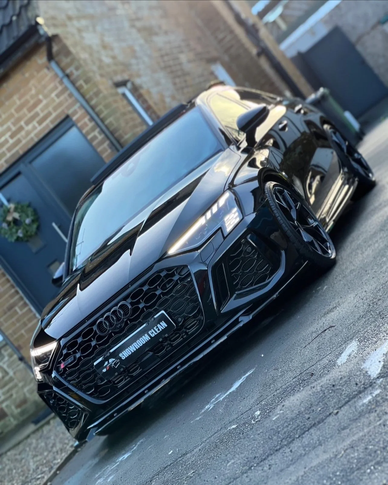 A black Audi sports car parked on a driveway outside a brick house with a blue door and a wreath, reflecting the sky and nearby surroundings.