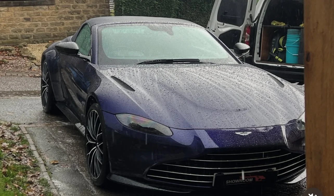 A dark blue convertible car with a grey roof parked on a wet asphalt driveway next to a brick wall and a wooden post, with raindrops on its surface.