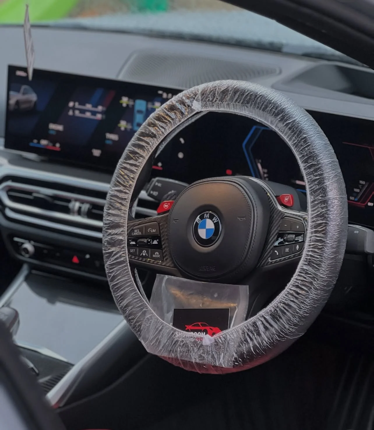 Interior of a BMW car showing a steering wheel wrapped in protective plastic, an electronic dashboard display, and various controls.