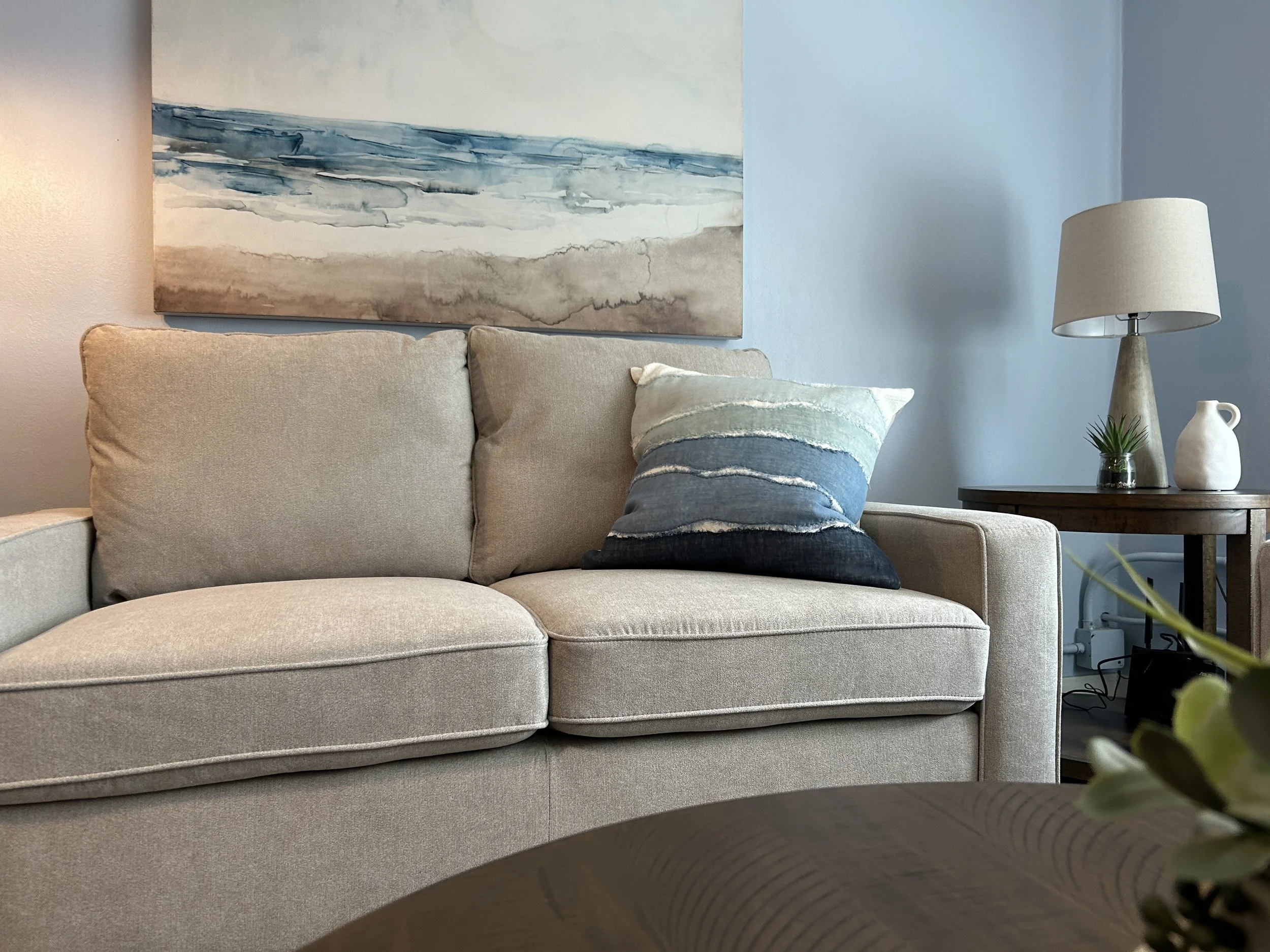 A beige sofa with pillows, a large abstract painting of a seascape hanging above it, a wooden side table with a lamp, a small potted plant, and decorative vases.