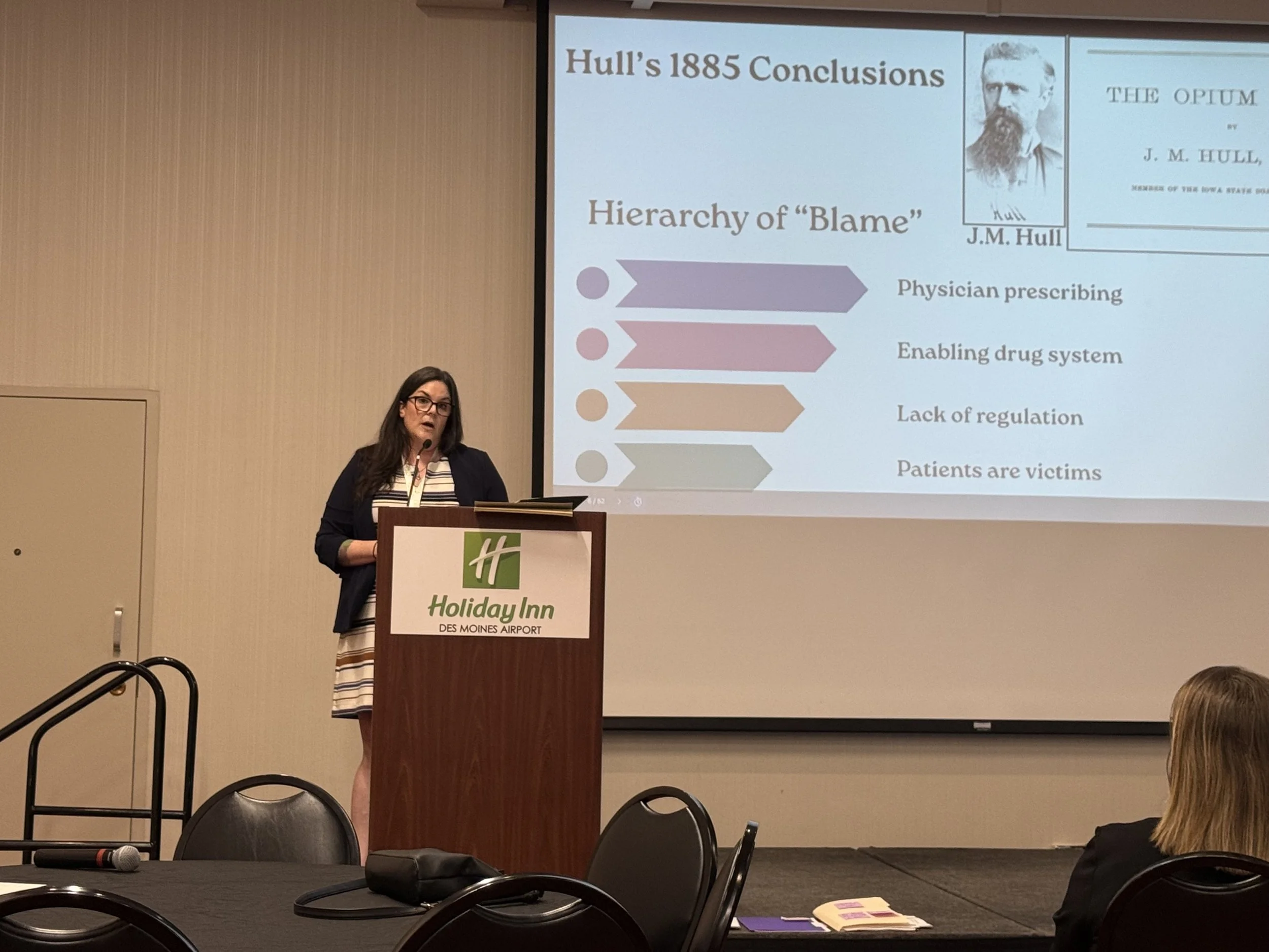 A woman with long dark hair and glasses standing at a podium labeled Holiday Inn, giving a presentation. A large screen behind her displays a slide titled 'Hull's 1885 Conclusions' with bullet points and images.