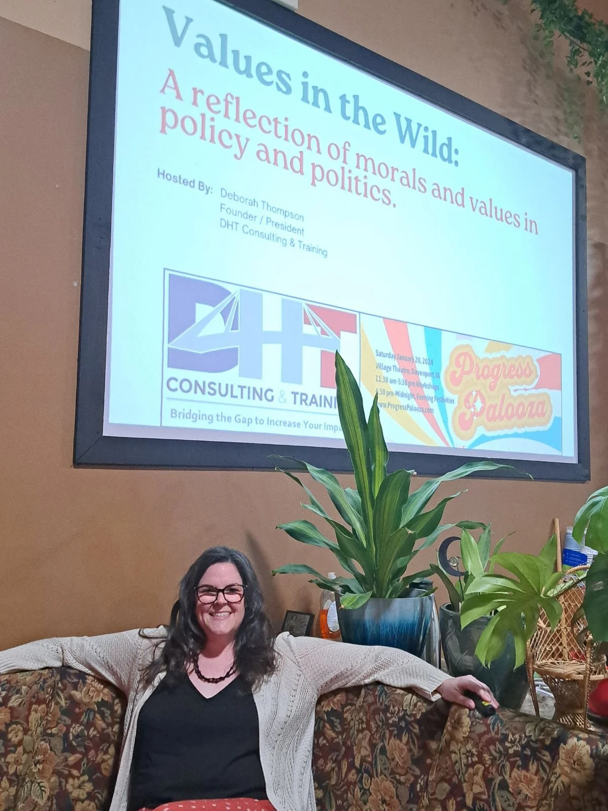 A woman with glasses and long hair smiling, sitting on a floral-patterned couch with her arm resting on the back. Behind her is a large potted plant, and a projection screen displaying a presentation titled "Values in the Wild: A reflection of morals and values in policy and politics." The presentation is hosted by Deborah Thompson, Founder/President of DHT Consulting & Training, at a venue named Progressive Palooza, with details of a workshop event on January 20, 2024.