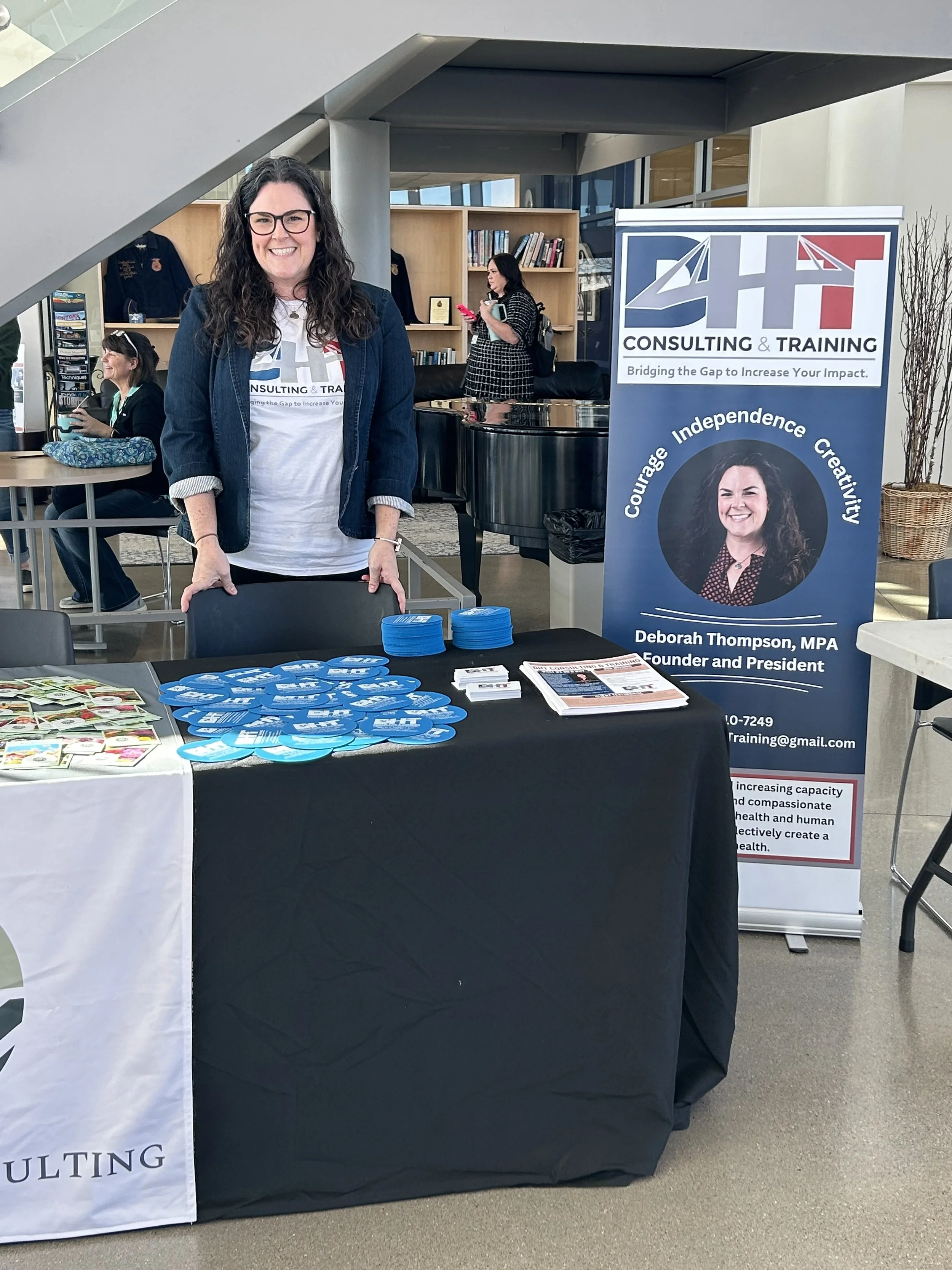 A woman with glasses and curly hair smiling at an indoor event, standing behind a table with campaign materials including buttons and brochures. A banner next to her reads 'C4H Consulting & Training' with a photo and information about Deborah Thompson, MPA, Founder and President. In the background, two women are seated and talking, with bookshelves and a piano visible.