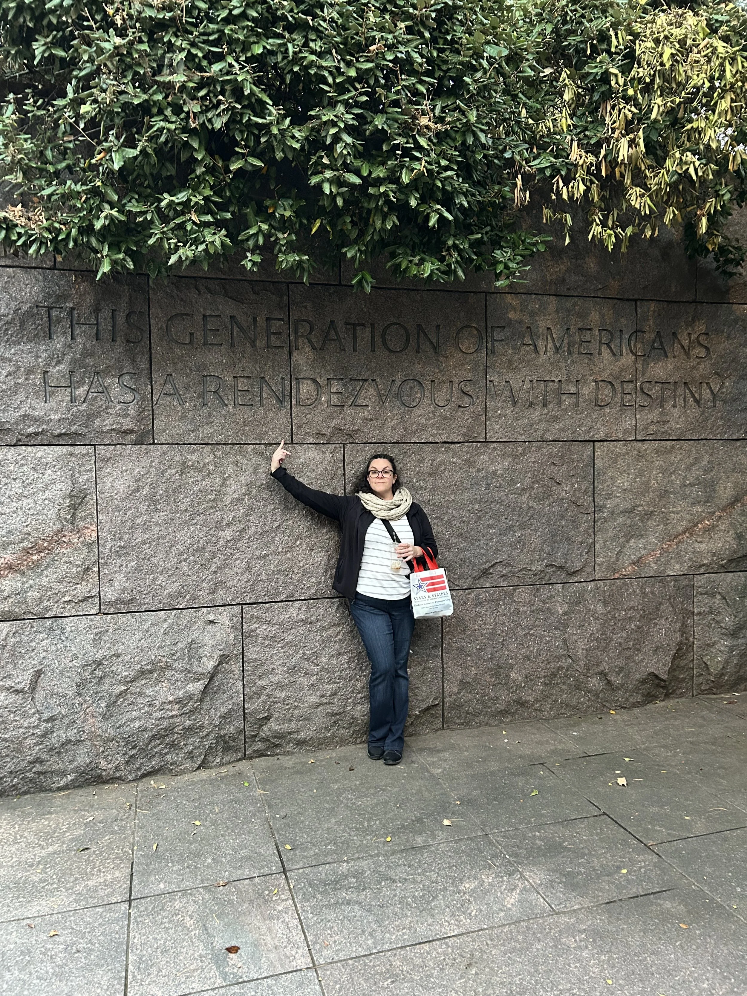 A woman standing against a large stone wall with an inscription that reads, 'This generation of Americans has a rendezvous with destiny.' She is wearing a black jacket, white and gray striped shirt, blue jeans, and black shoes. She has glasses, a scarf, and is holding a drink in one hand and a bag with American flag and star designs over her shoulder. She is pointing at the inscription in the wall.