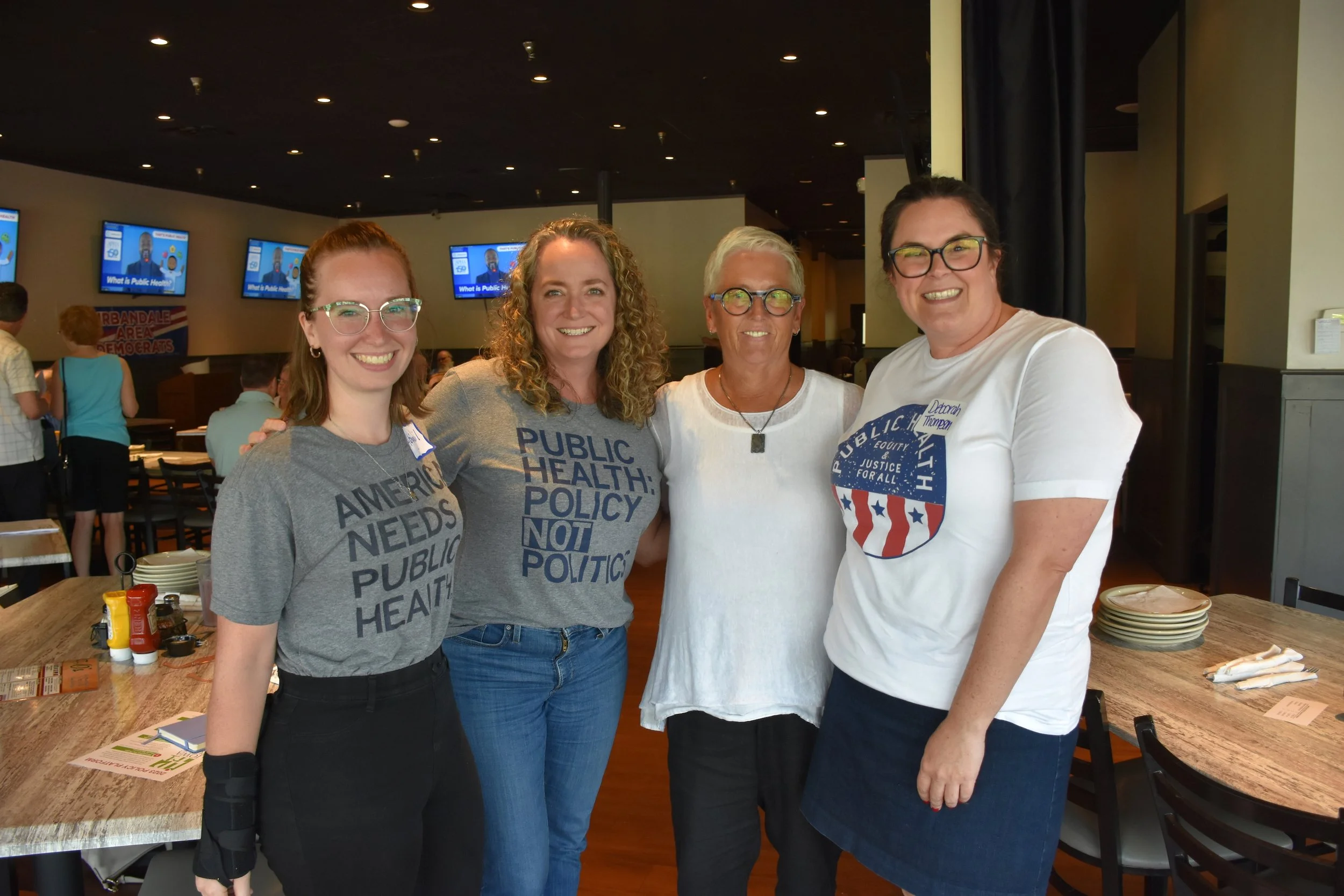 Four women standing together inside a restaurant, smiling at the camera. Three of them are wearing gray and white T-shirts with political slogans related to public health and policy. In the background, there are multiple television screens and a group of people near a sign that reads 'URBANDALE AREA DEMOCRATS.'