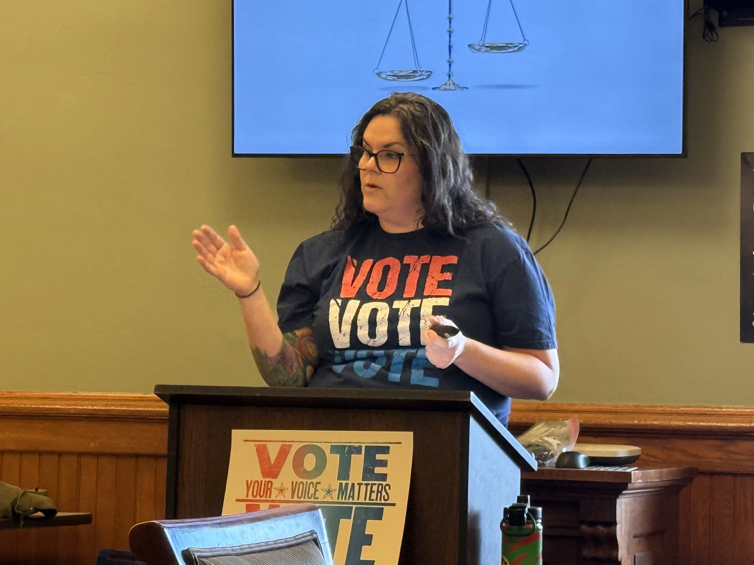 A woman with dark curly hair, glasses, and tattoos on her arm is standing behind a podium with a sign that reads 'VOTE YOUR VOICE MATTERS' in front of a flat screen tv showing a drawing of a balance scale. She appears to be speaking or presenting in a room with wood paneling and beige walls.