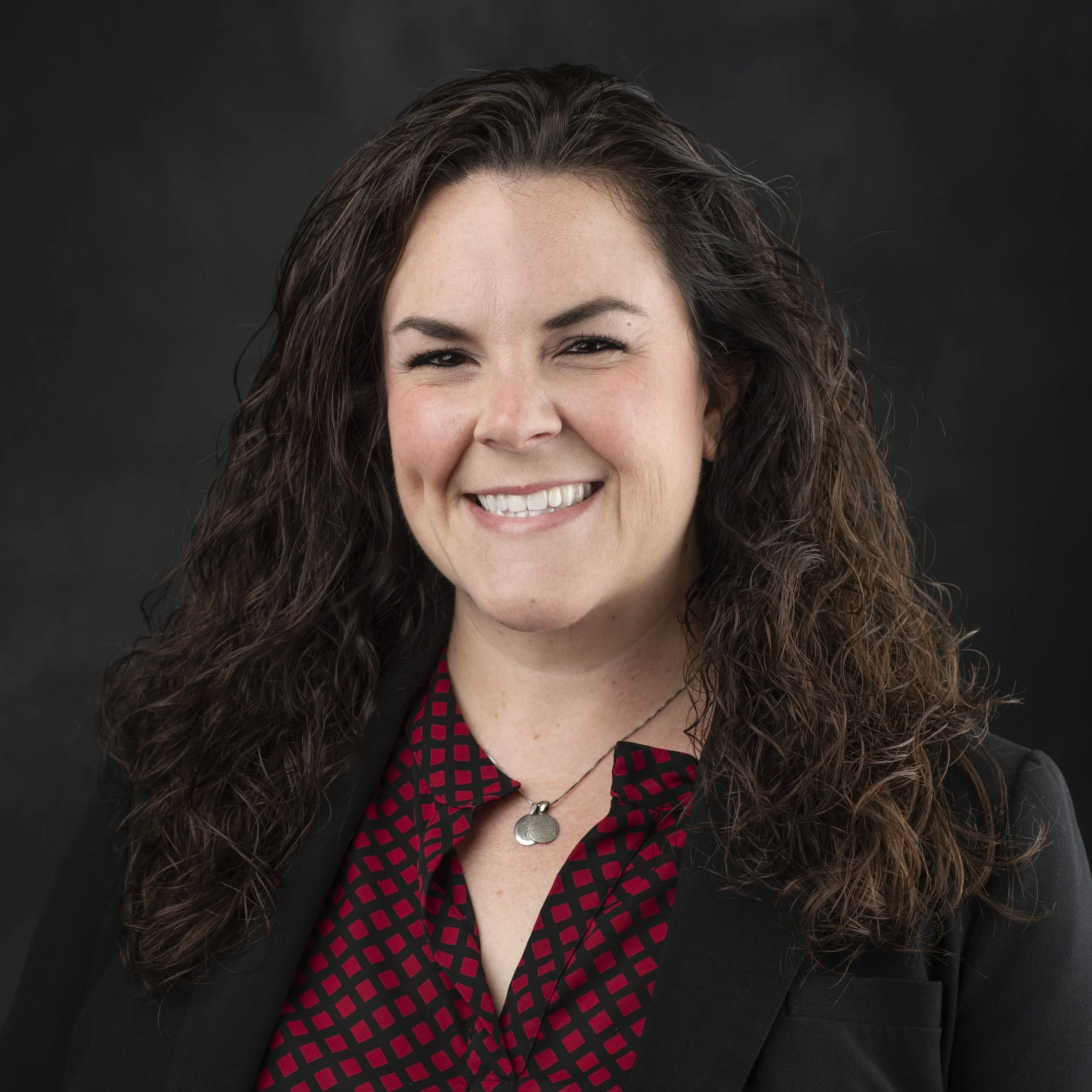 A woman with long curly brown hair smiling and showing her teeth, wearing a black blazer and a red and black checkered shirt underneath, with a silver necklace and a large round pendant, against a dark background.