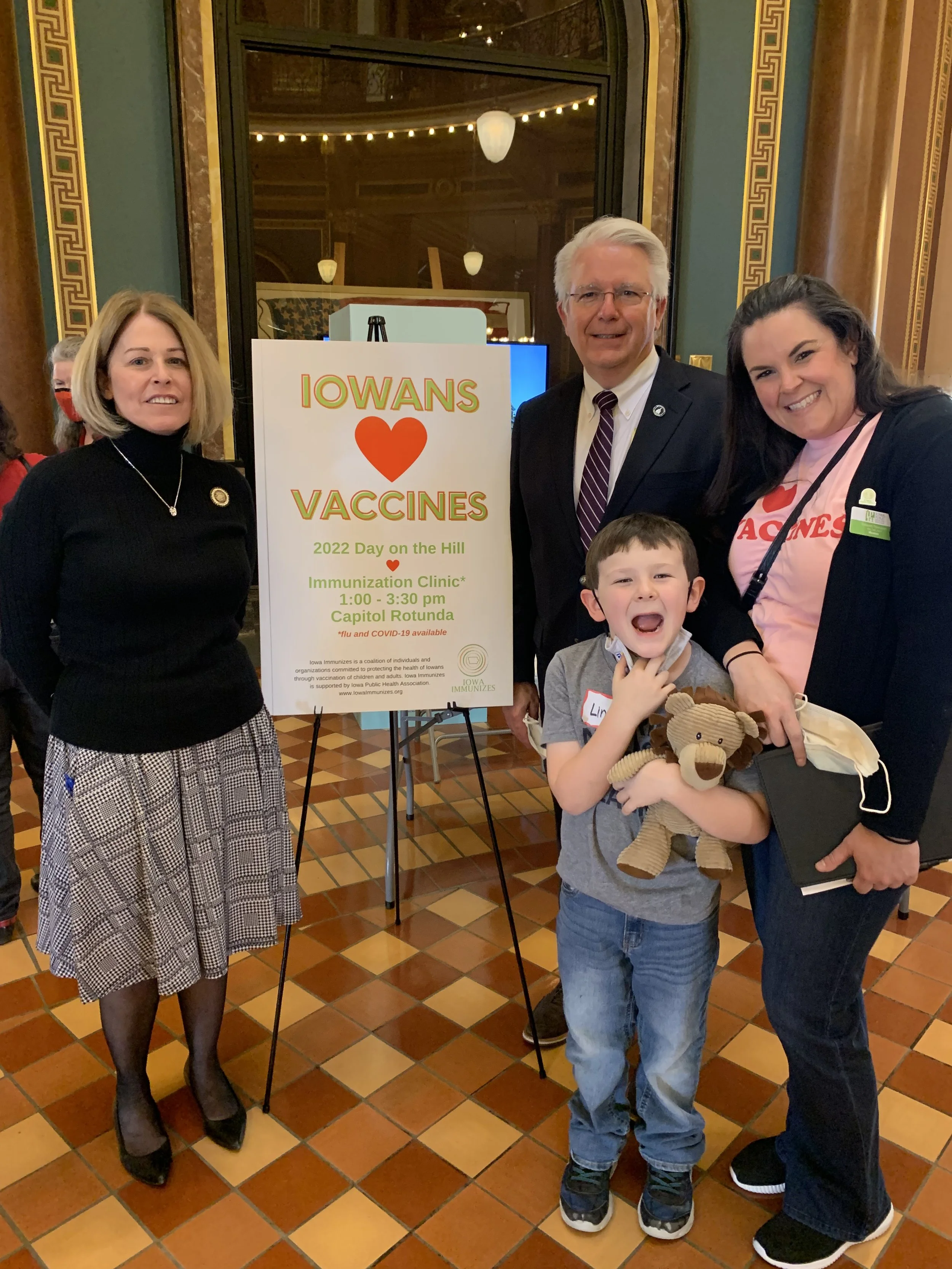 A group of four people, three adults and one child, standing in front of a sign at a vaccine event. The sign reads 'IOWANS ❤️ VACCINES,' with details about a 2022 immunization clinic. The adults are smiling, and the child is holding a teddy bear and has a wide, open-mouthed smile.