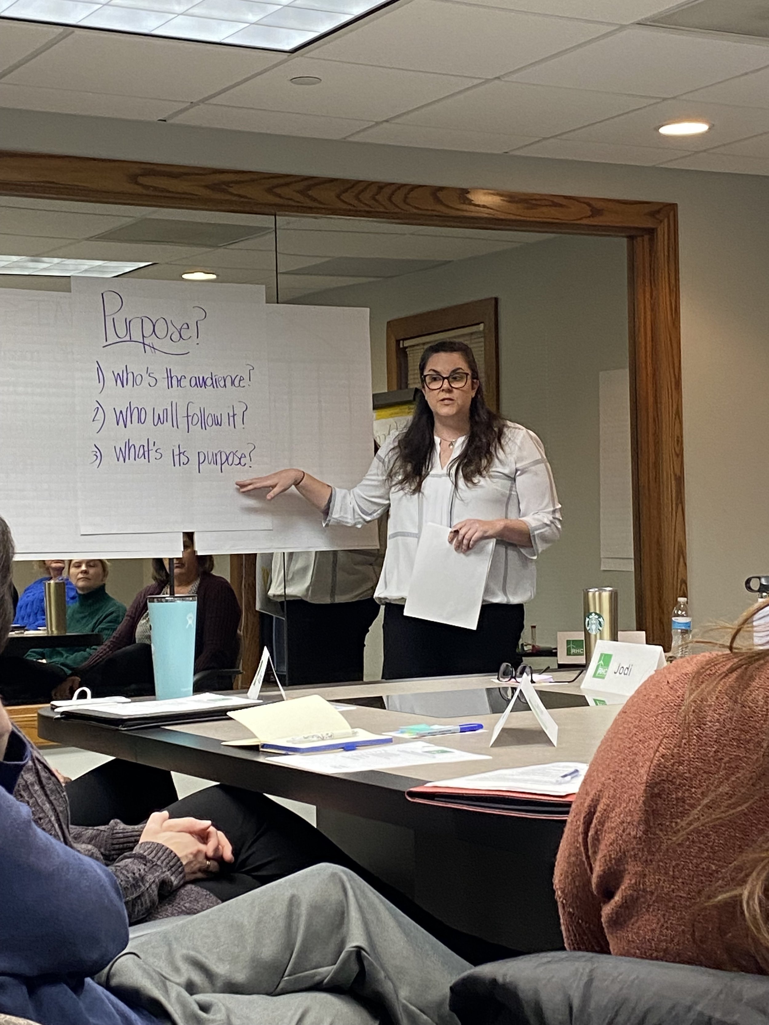 A woman in glasses and a white shirt is standing in front of a whiteboard, giving a presentation to an audience in a conference room. The whiteboard has handwritten notes about purpose. Several people are seated at a long table, listening attentively, with some taking notes and papers on the table.