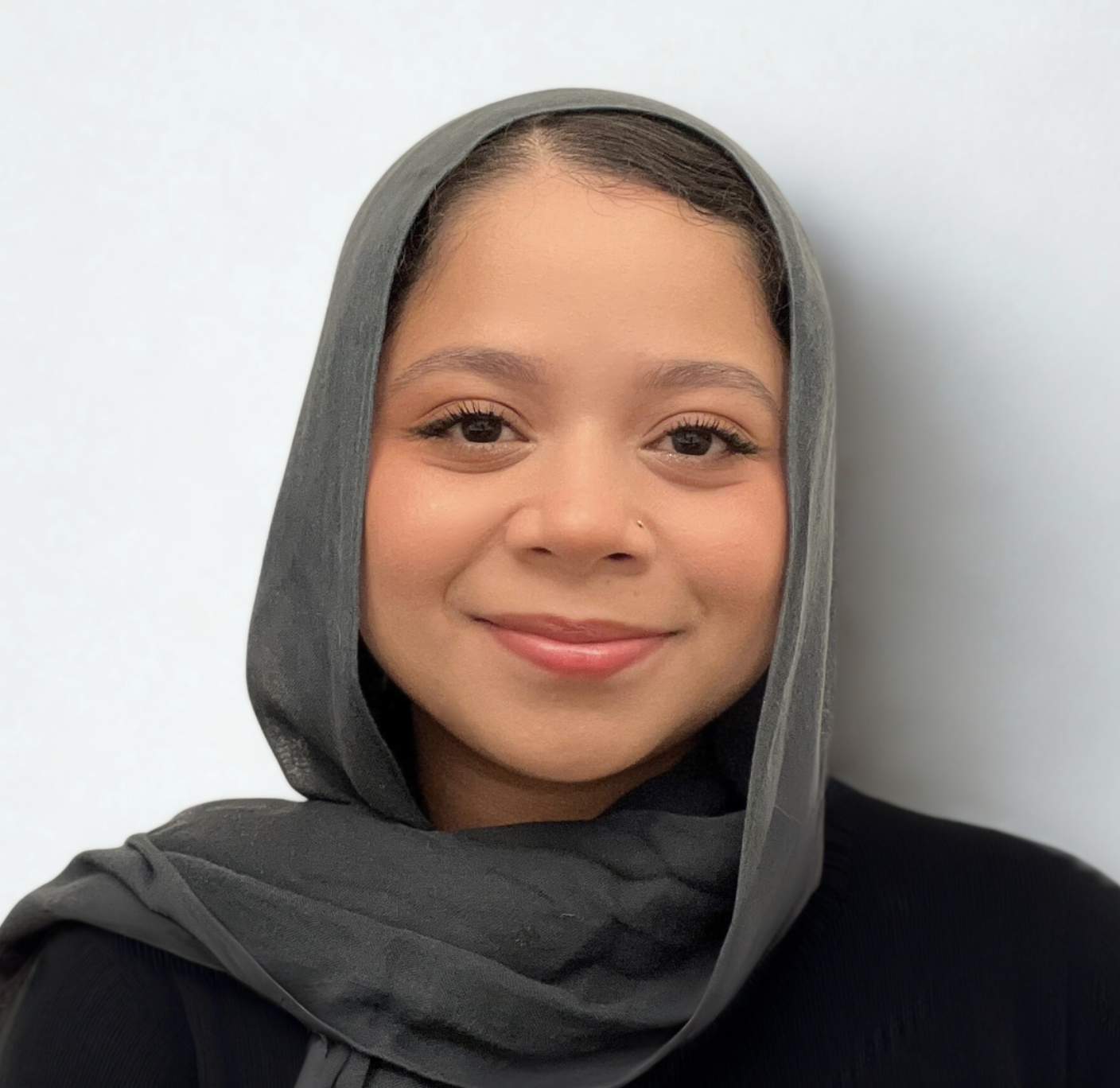 A smiling woman with black hair, wearing a headscarf and black sweater, standing against a white background.