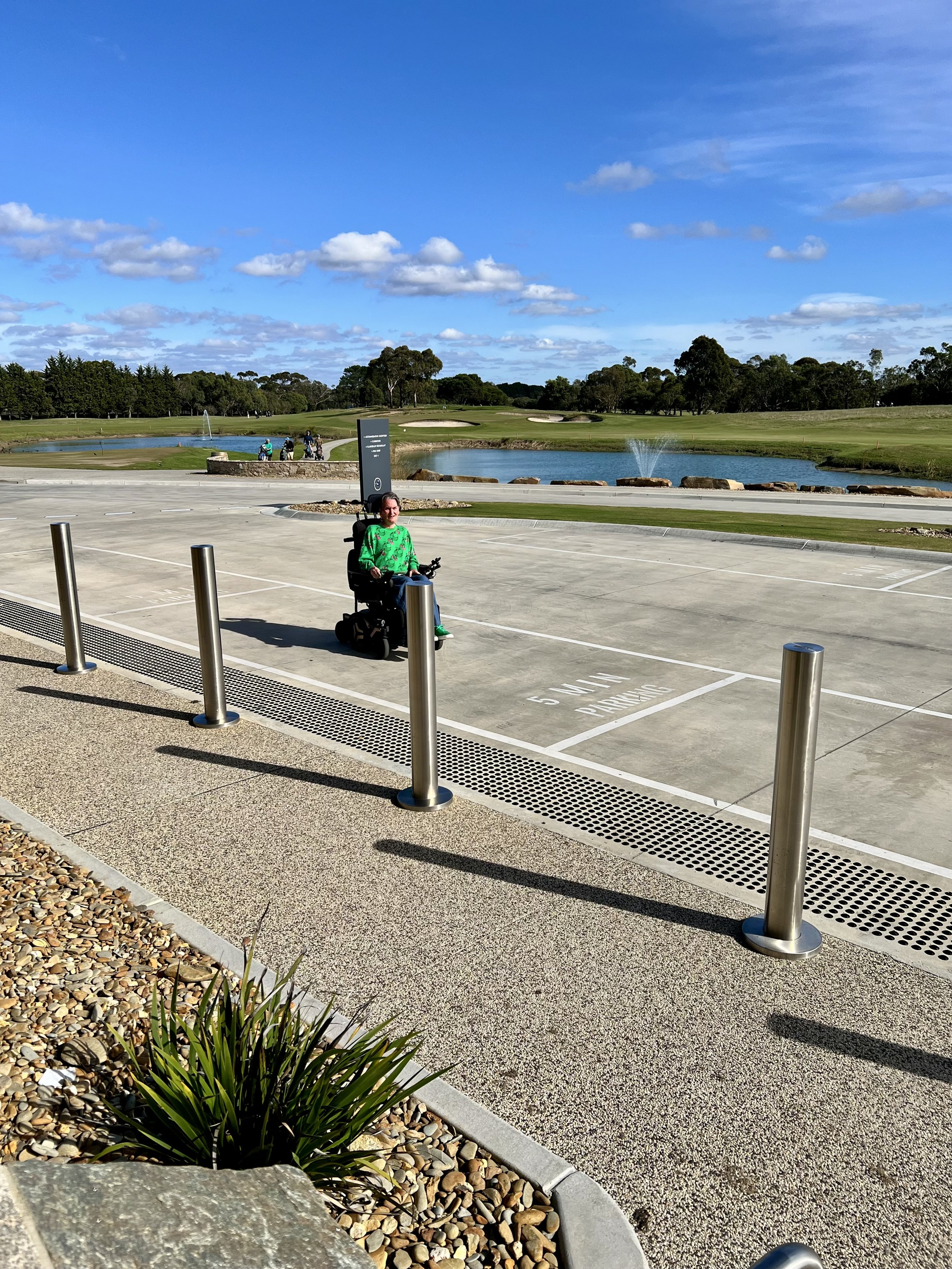  Blue sunny sky with white clouds dark green Bush like treeswith golf course Green ground with a small blue lake  in the background . Woman in a bright green top sitting in a wheelchair on the wide pale concrete drive there are silver bollards in fro