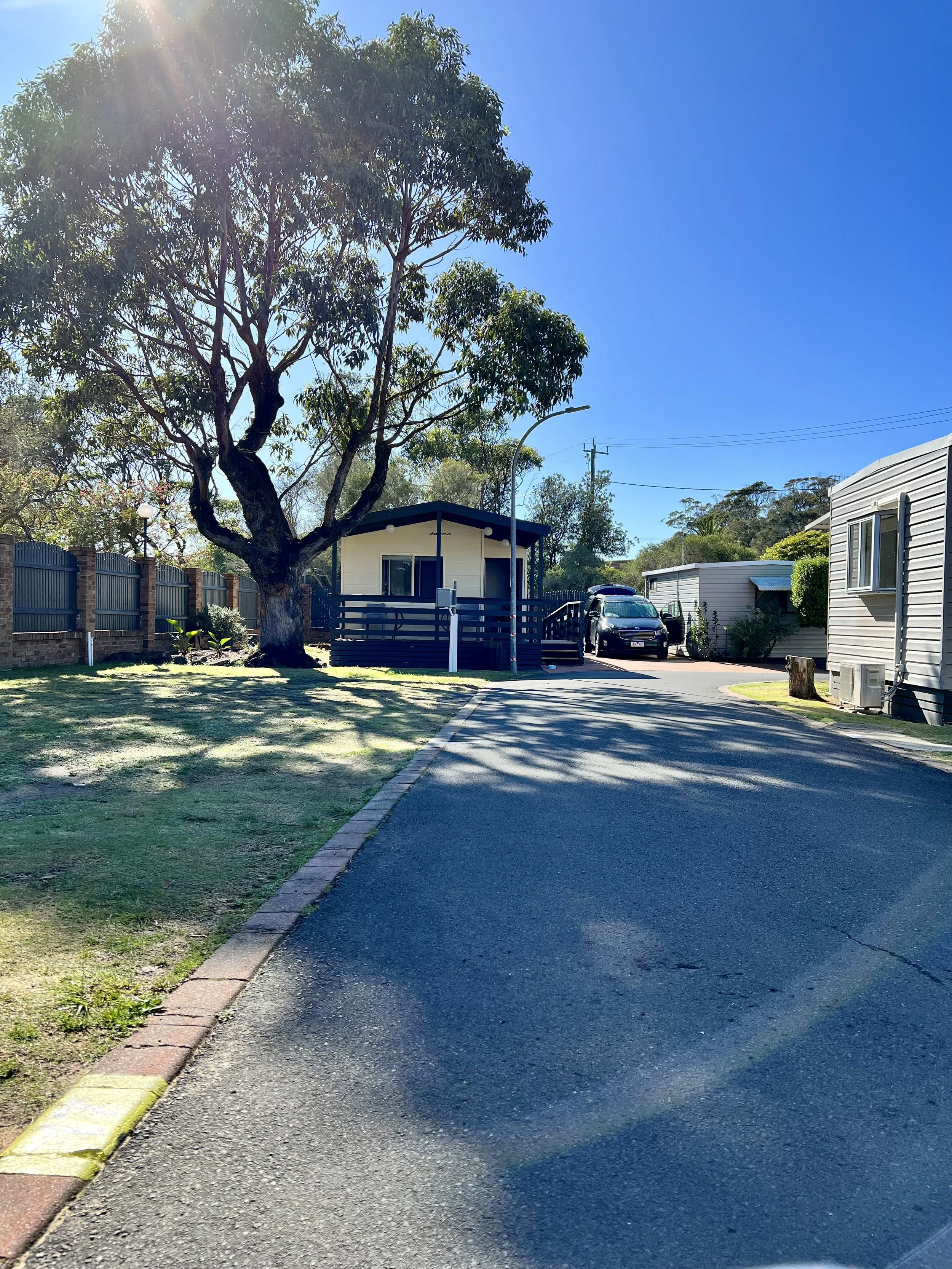 Looking at the front of the Cream weatherboard and Brown eaves and railing of the wheelchair accessible unit. Four steps are shown to the right of the Veranda with a doorway and one window. There is a car reversed into the space beside the unit