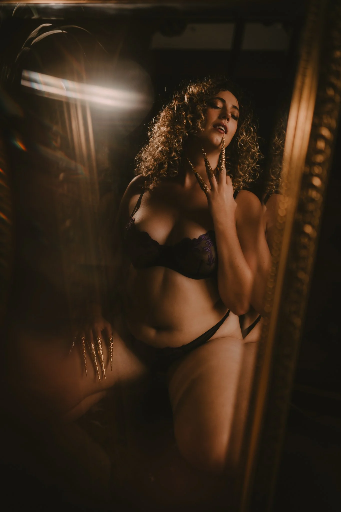 A woman with curly hair wearing black lingerie and gold jewelry, posing in front of a mirror with warm lighting, creating a sensual atmosphere.