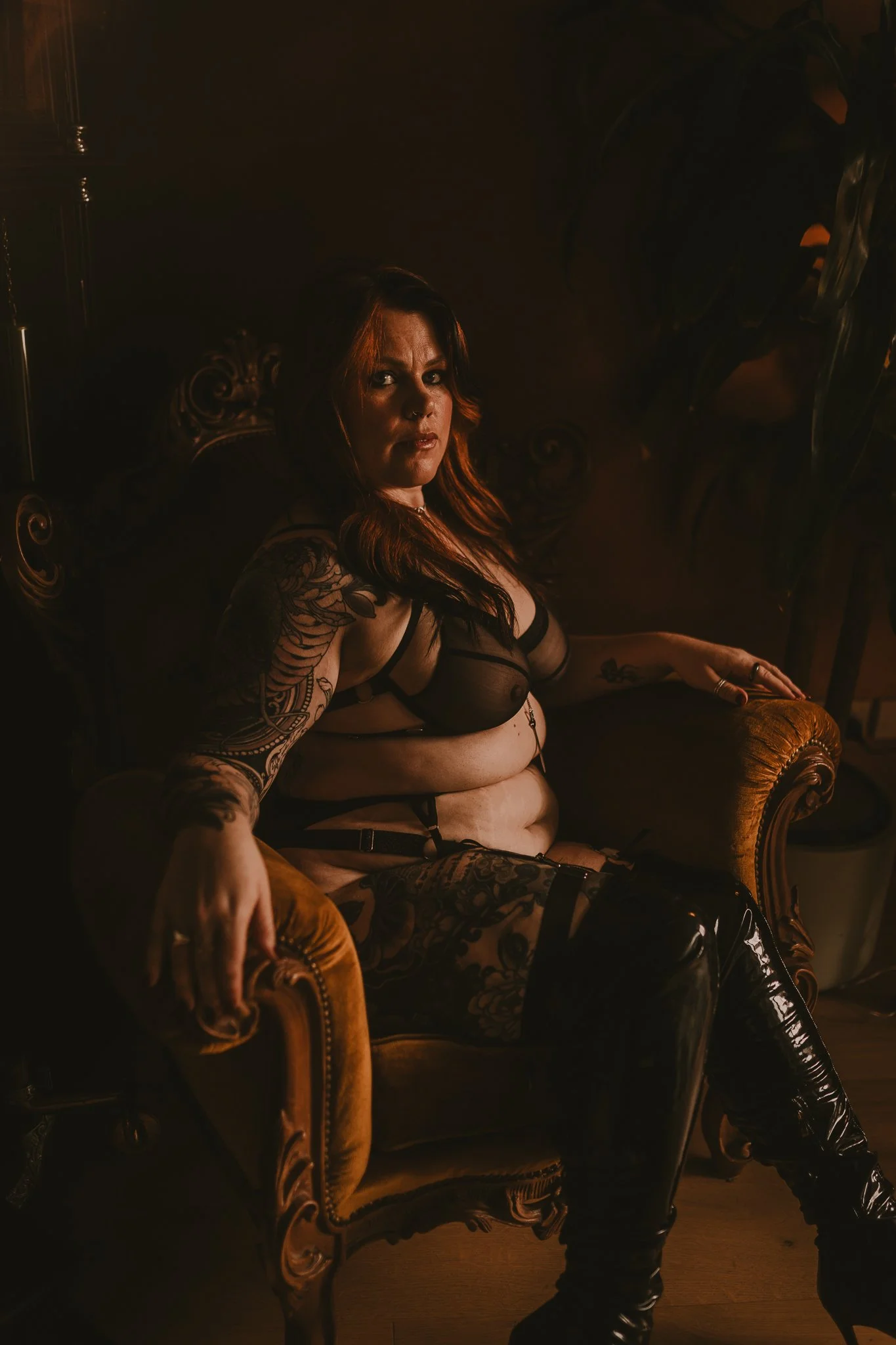 A woman with long, dark hair and tattoos sitting on an ornate, vintage armchair, wearing black mesh lingerie and thigh-high patent leather boots, in a dimly lit room with warm, moody lighting.