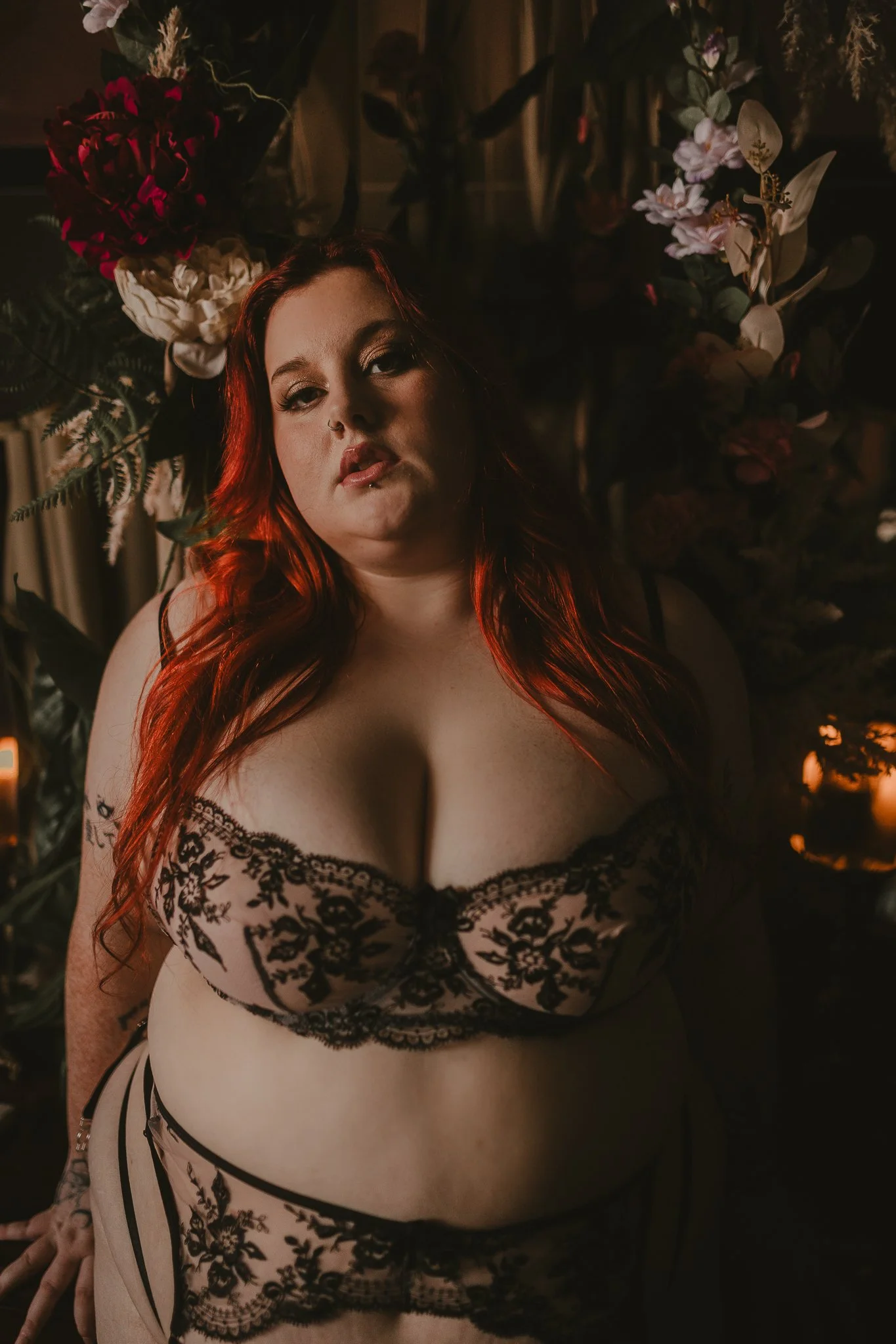 A woman with long red hair wearing black lace lingerie in a dimly lit room with floral arrangements in the background.