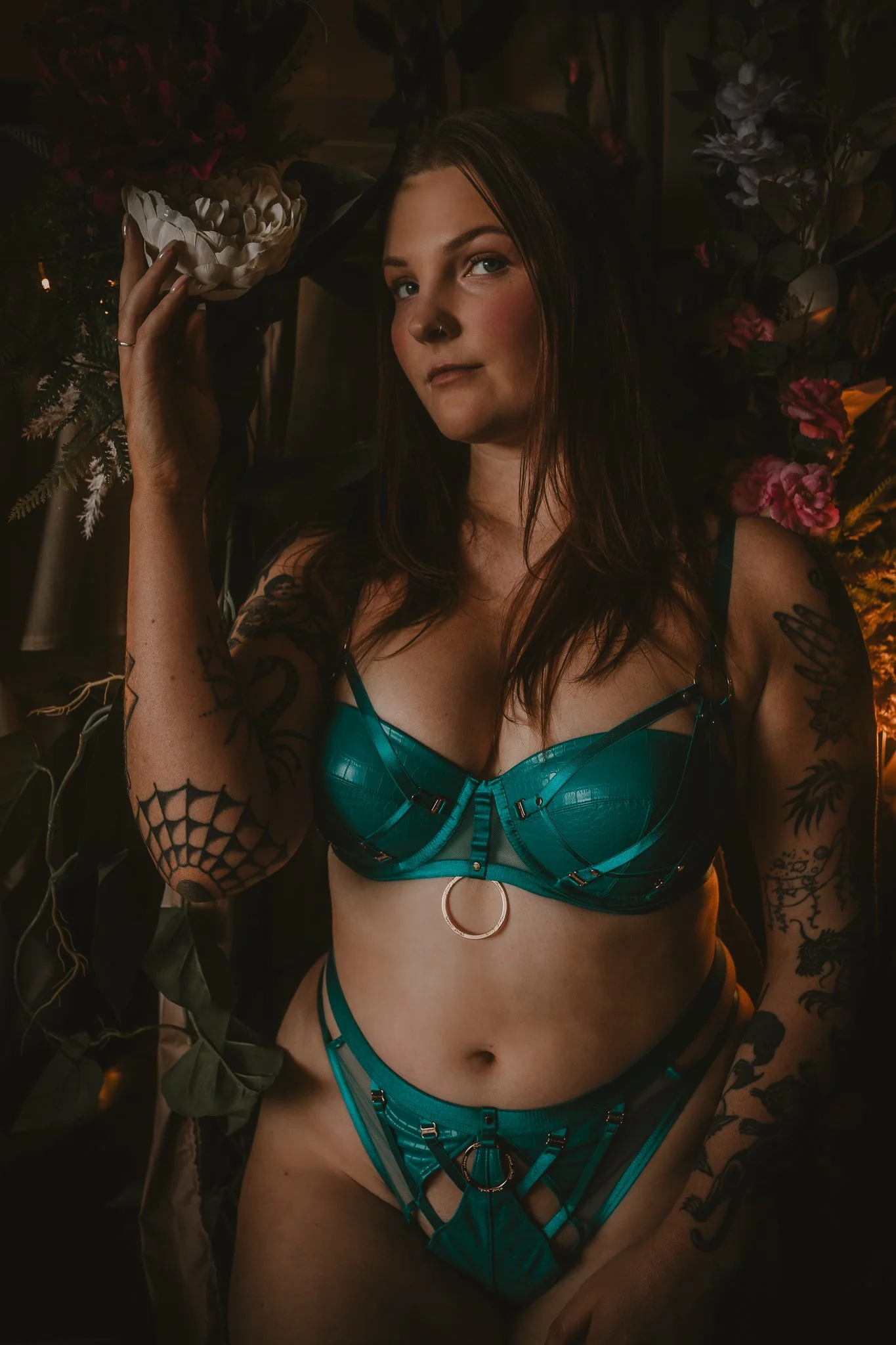 A woman with long brown hair and tattoos on her arms and shoulder, wearing teal lingerie with harness details, standing among flowers and plants, holding a flower close to her face.