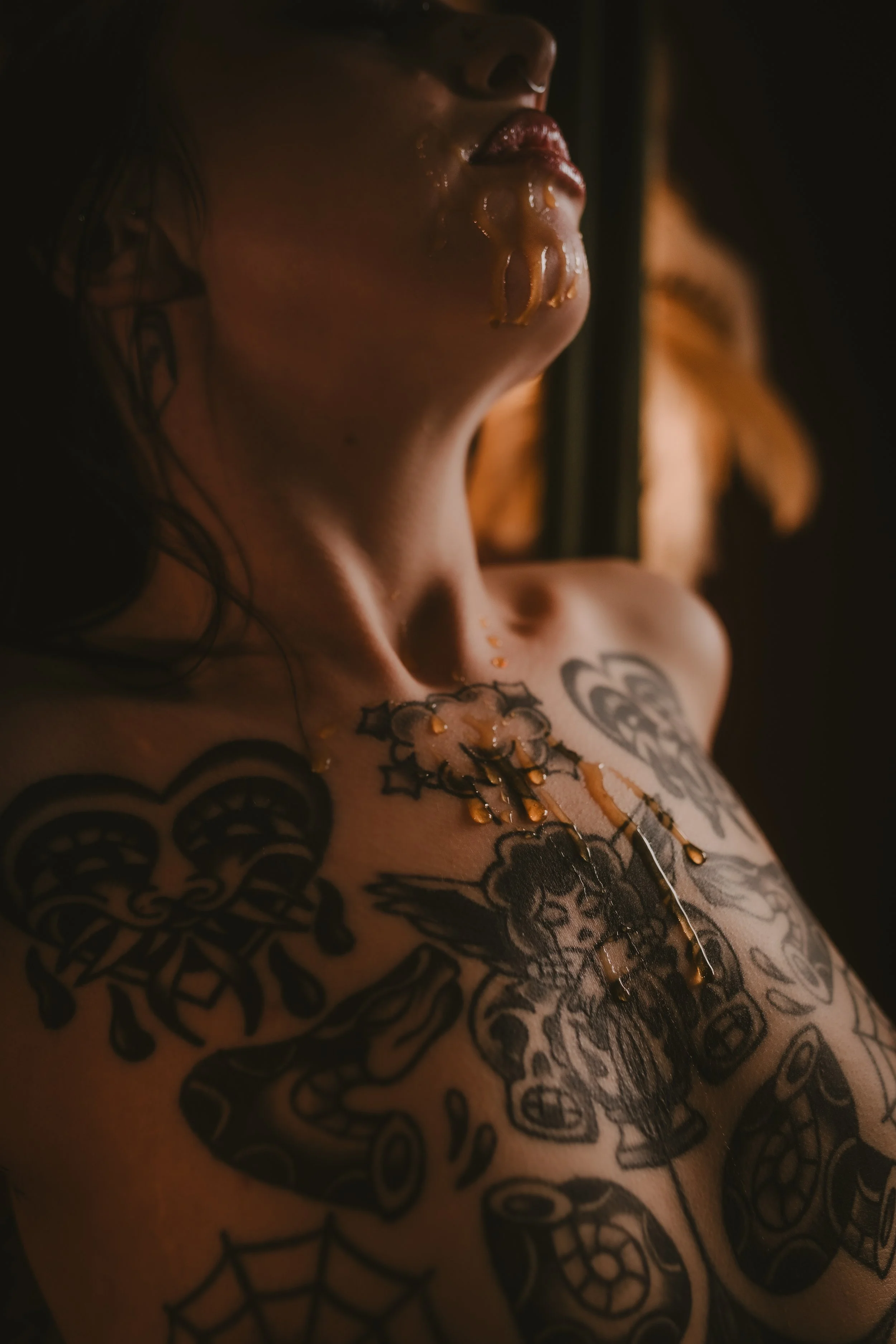 Close-up of a person with tattoos on their chest, neck, and face, with honey or syrup dripping from their lips and chin in a dark setting.