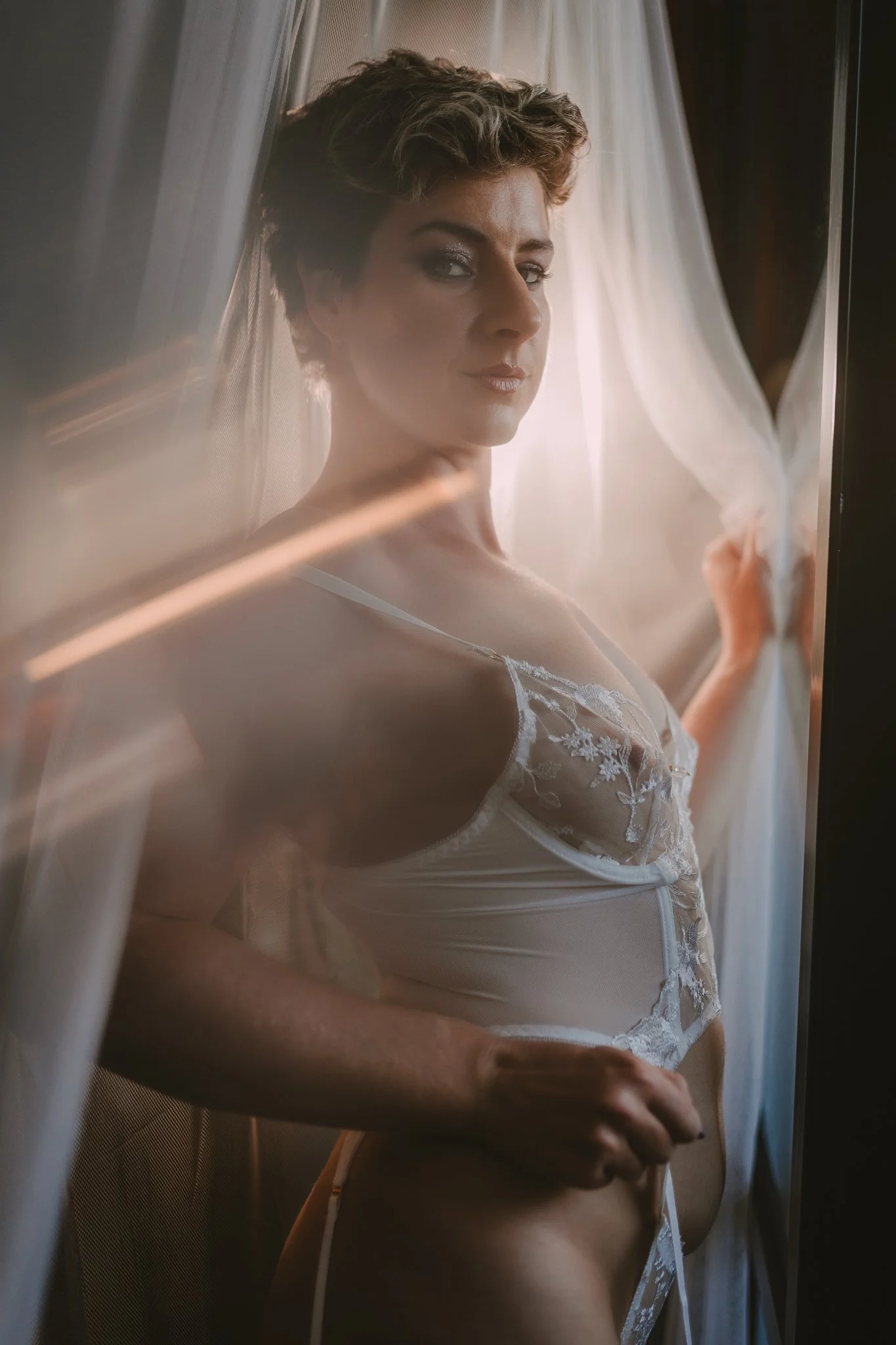 A woman with short, styled hair and makeup wears white lace lingerie and stands behind sheer curtains, looking at the camera with a confident expression.