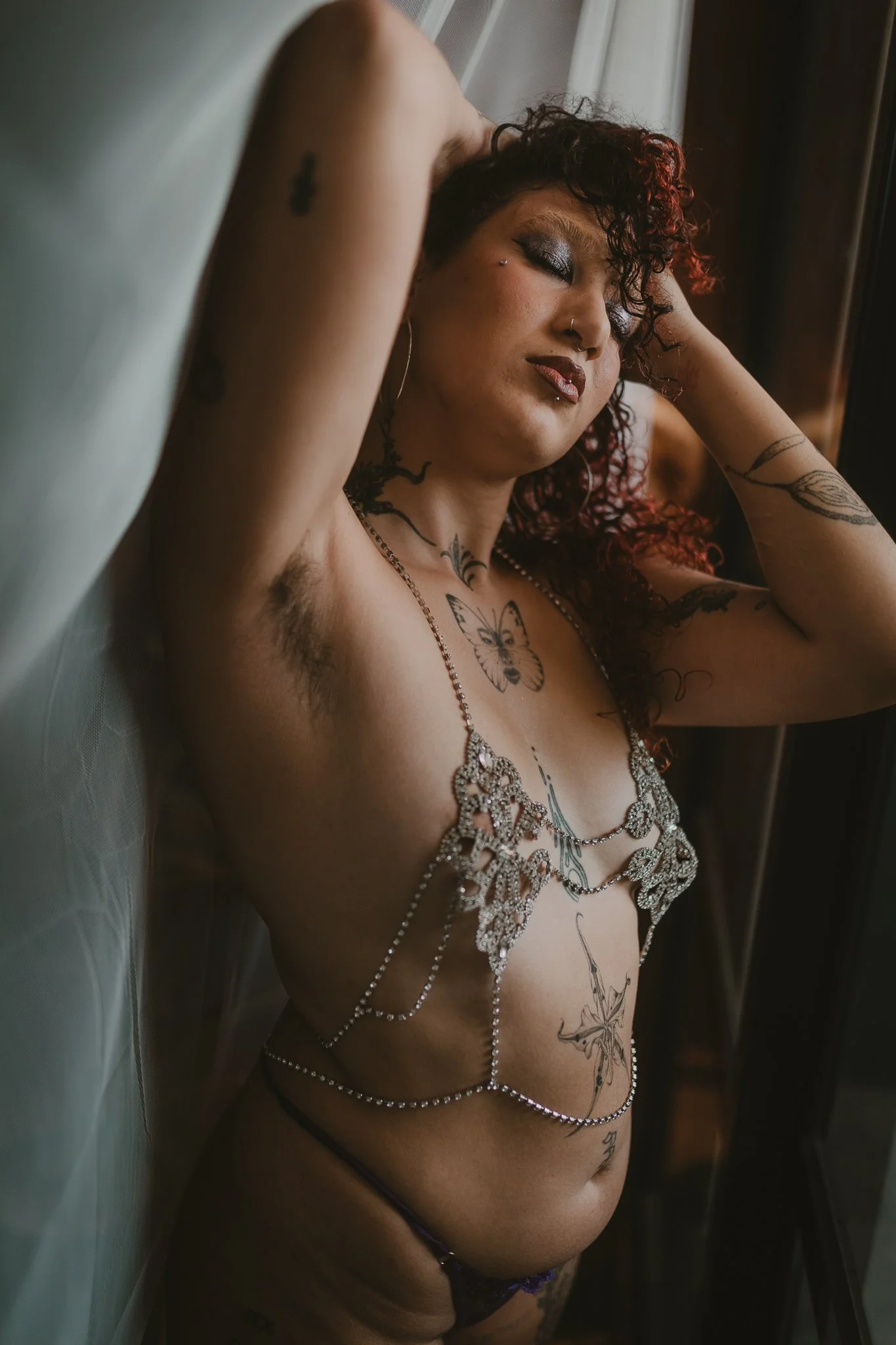 A woman with tattoos and dark curly hair, wearing jewelry, relaxing with arms behind her head near a window
