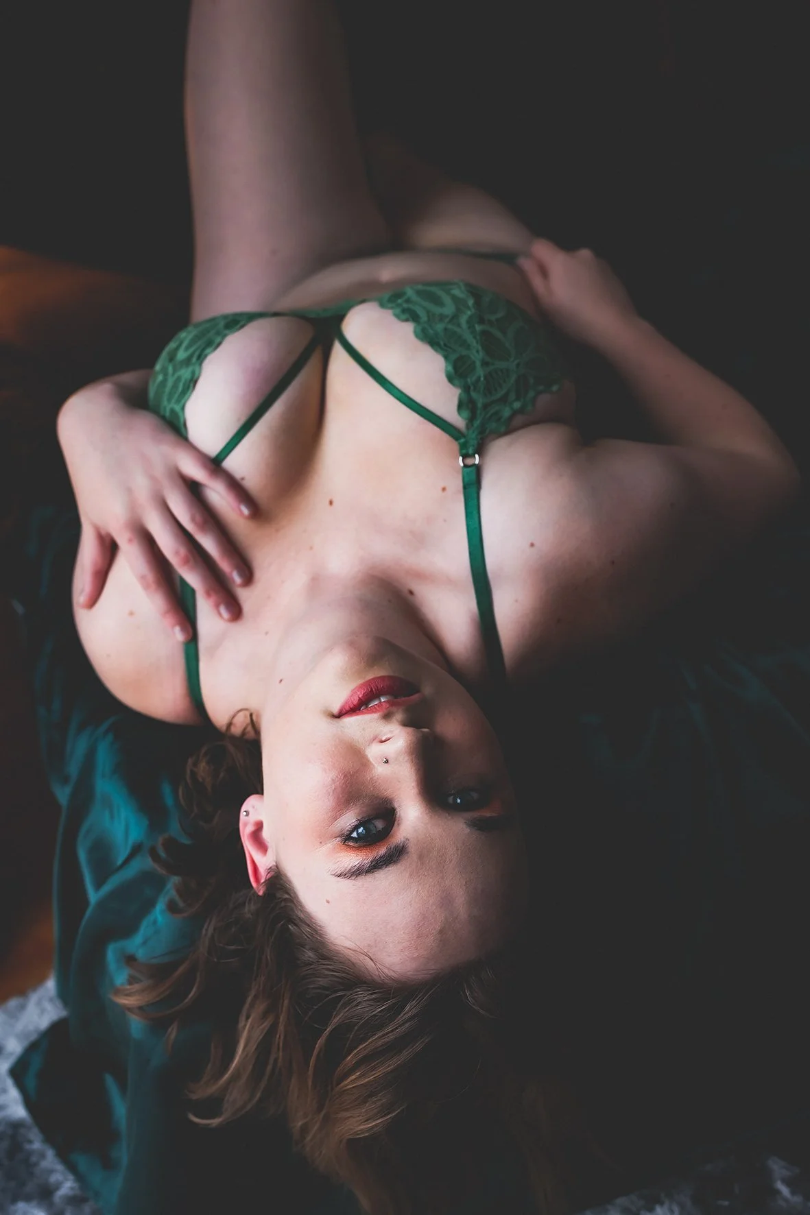 A woman lying on her back on a dark surface, wearing green lace lingerie, looking up at the camera with a relaxed expression.