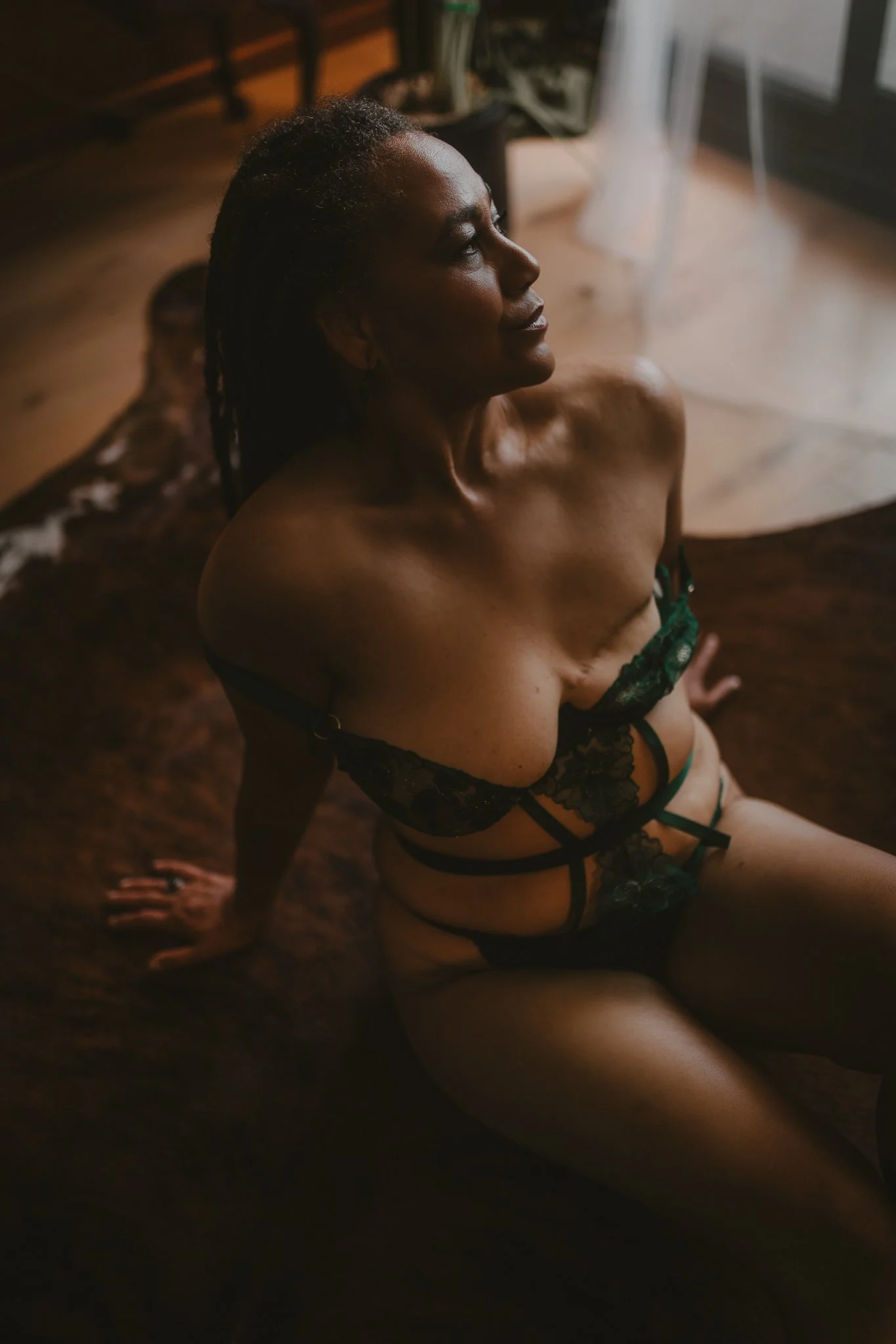 A woman with dark skin and dreadlocks wearing black lace lingerie sitting on a brown rug in a warmly lit room.